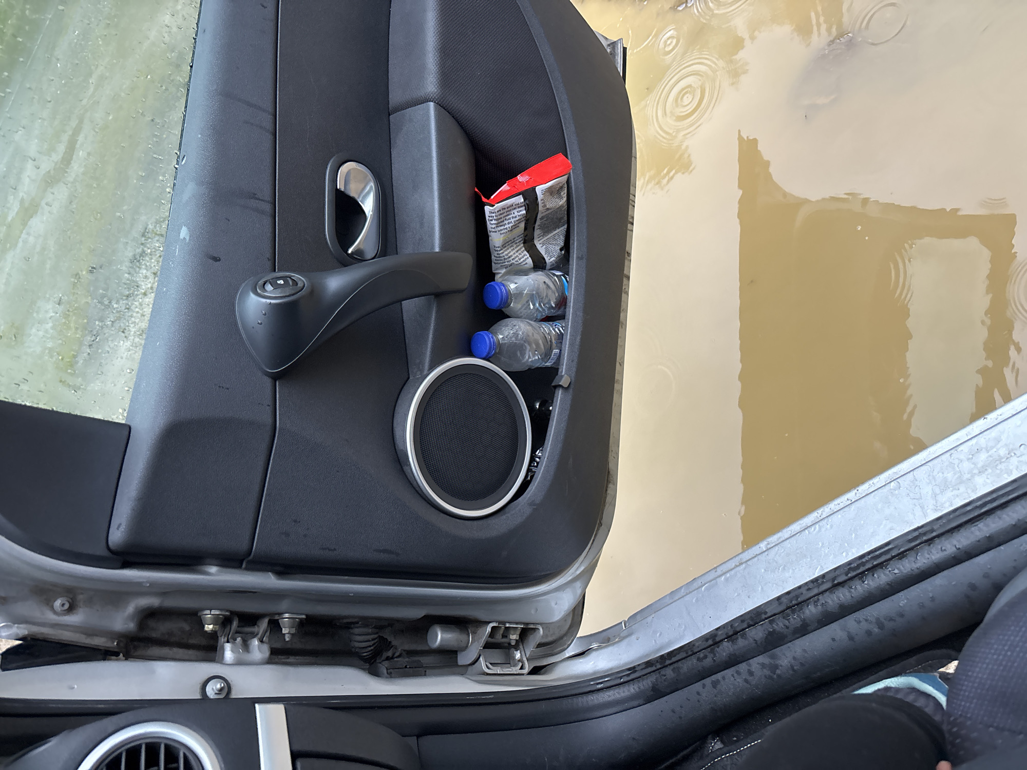 The image shows the interior of a car door in what appears to be a flooded area. The car door panel is black plastic with a speaker, door handle, and a storage compartment. Inside the storage compartment are two plastic water bottles and a crumpled bag, possibly of chips or snacks. The exterior of the car door is silver. Outside the car, muddy water is visible with ripples indicating movement, suggesting rain or ongoing flooding. The surrounding environment is indistinct due to the water. The image is taken from a low angle, looking into the car. There are no people clearly visible in the scene, although a portion of a person's leg and foot in athletic footwear can be seen at the bottom left of the frame. The scene appears to be in an outdoor setting, possibly a street or a parking lot, given the presence of floodwater. The location is specified as Alamovtsi, Bulgaria.