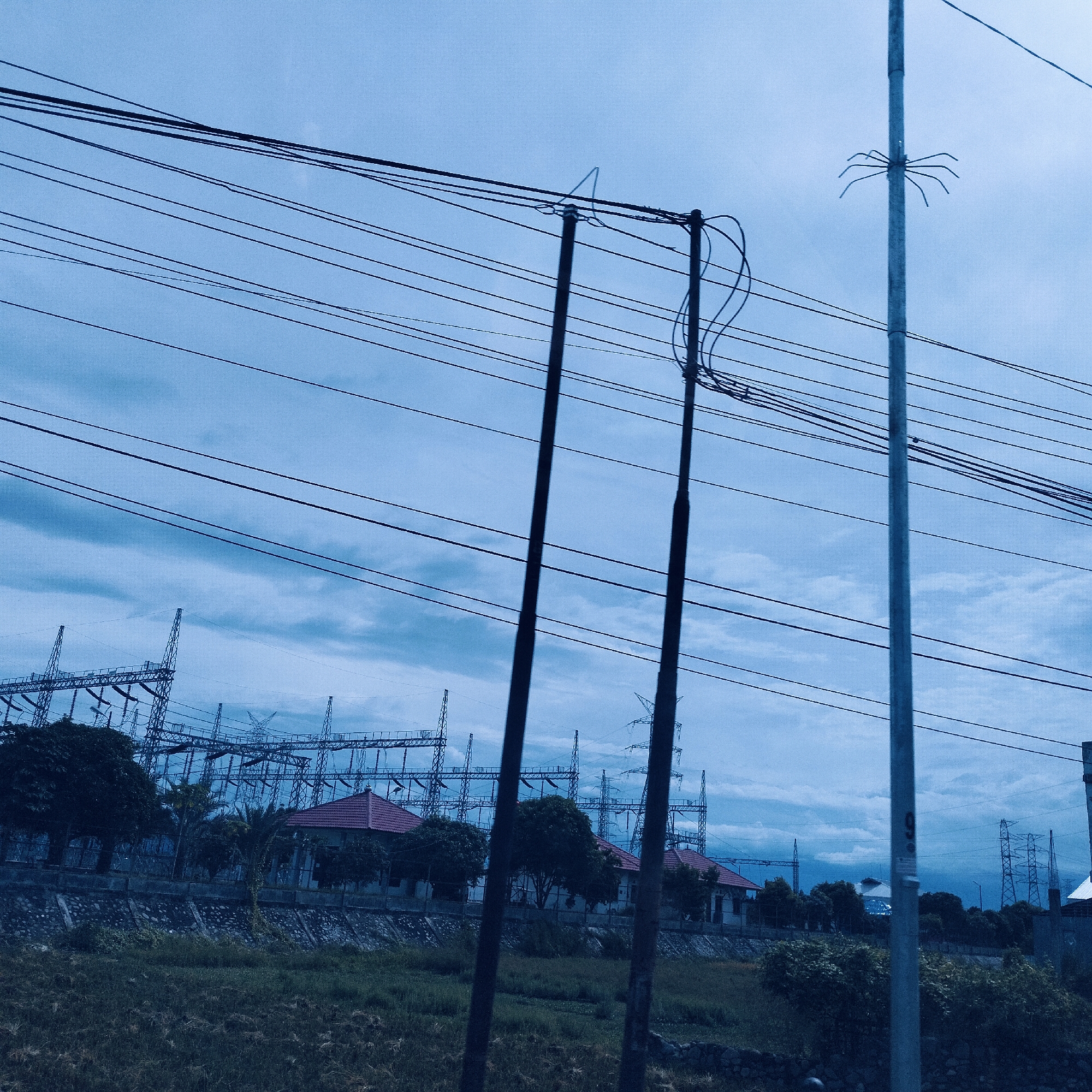 The image displays an outdoor scene dominated by electrical infrastructure under an overcast, blue-hued sky. In the foreground, numerous dark power lines stretch horizontally across the frame, supported by several utility poles. Two prominent dark poles stand near the center, with one showing coiled wires and another a triangular wire support. To the right, a lighter-colored pole features a distinct spider-like metal apparatus attached to it.
The midground shows a patch of green grass or field, bordered by a low concrete wall. Behind this wall, several dark-leaved trees are visible, along with multiple houses characterized by their distinctive red-tiled roofs. In the background, a large electricity substation or power plant extends across the horizon, consisting of a complex array of tall, metallic structures and transmission towers.
The overall color palette is cool and desaturated, indicative of either an overcast day or a specific photographic filter. The heavy cloud cover suggests a subdued, potentially rainy or pre-rainy atmosphere. The scene is static, with no human presence or activities visible. The setting points to an industrialized or semi-urban area, consistent with Teungoh Baro, Indonesia. No prominent text is visible within the main image.