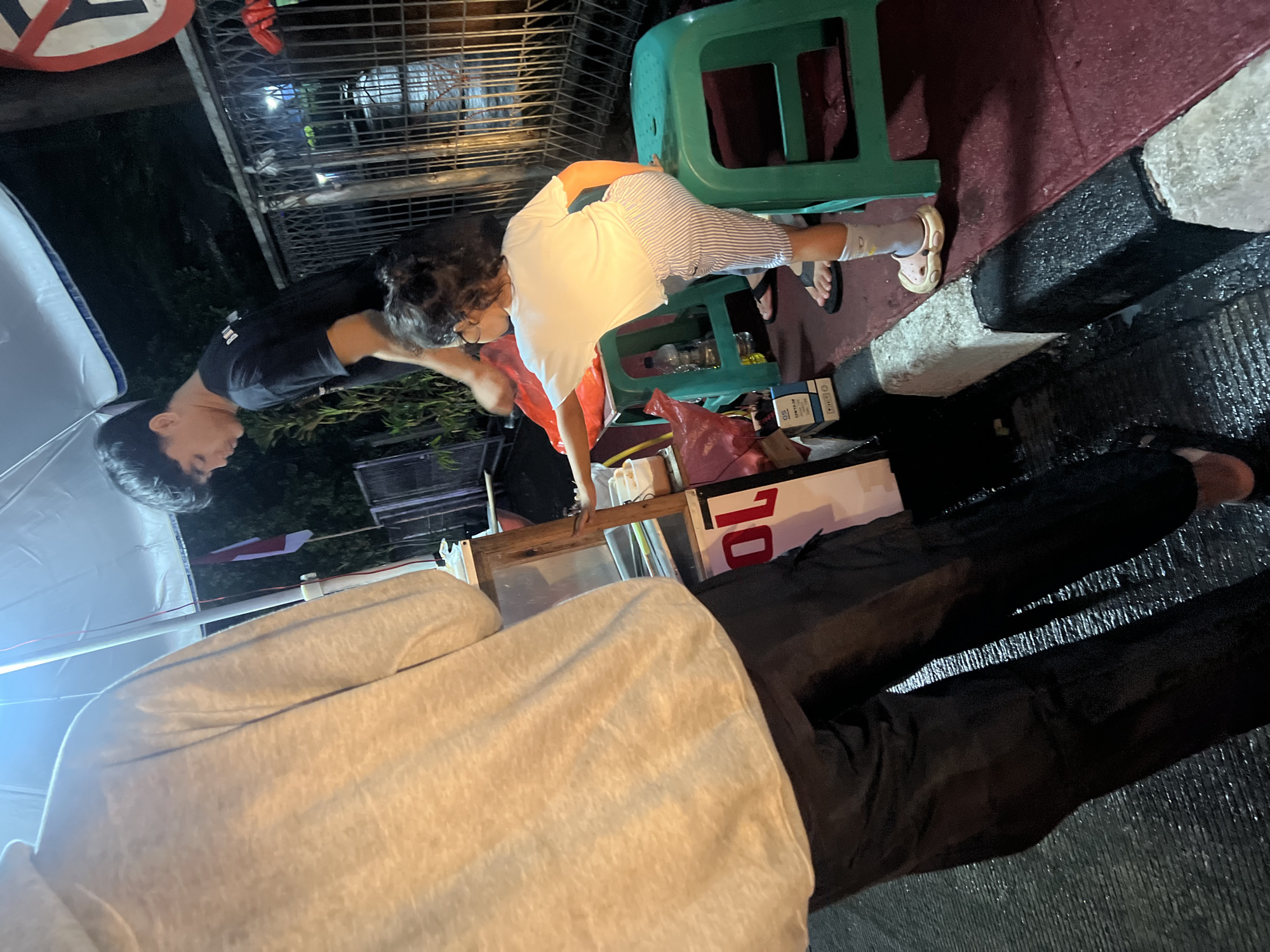 A young child, wearing a white t-shirt and striped pants, sits on a green plastic stool beside a food cart. The child is reaching towards the cart, possibly interacting with the vendor or the items on display. Behind the child, a young man in a black t-shirt is attending to the cart. To the left, a person wearing a light-colored hoodie and dark pants stands facing away from the camera, partially obscuring the food cart. The scene appears to be outdoors at night, with artificial lighting illuminating the area. The food cart has a wooden frame and what appears to be a white umbrella overhead. A red and white flag, likely the Indonesian flag, is visible in the background. The ground is wet, suggesting recent rain. The overall setting suggests a street food vendor operating in a public area. The location is identified as Gunung Putri, Indonesia.