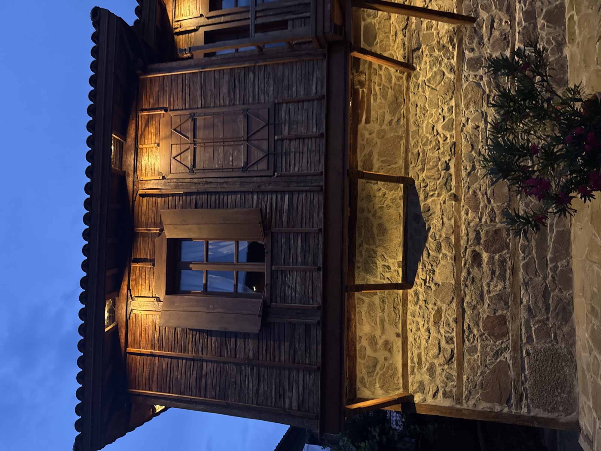 The image depicts the facade of a traditional wooden house in Sozopol, Bulgaria, illuminated by warm lights at dusk. The upper story is constructed with dark wooden planks, featuring two windows with open wooden shutters. One window shows a glimpse of curtains and interior lighting, while the other has decorative wooden lattice work over its closed shutter. The roof is covered with dark, curved tiles. Below the wooden story, the house has a stone foundation, built with irregularly shaped stones of varying colors. Wooden beams support the overhang of the upper story. In the foreground, a potted plant with dark green leaves and small pink flowers is visible. The sky above is a deep blue, indicating evening twilight. The overall scene conveys a historic and tranquil atmosphere.