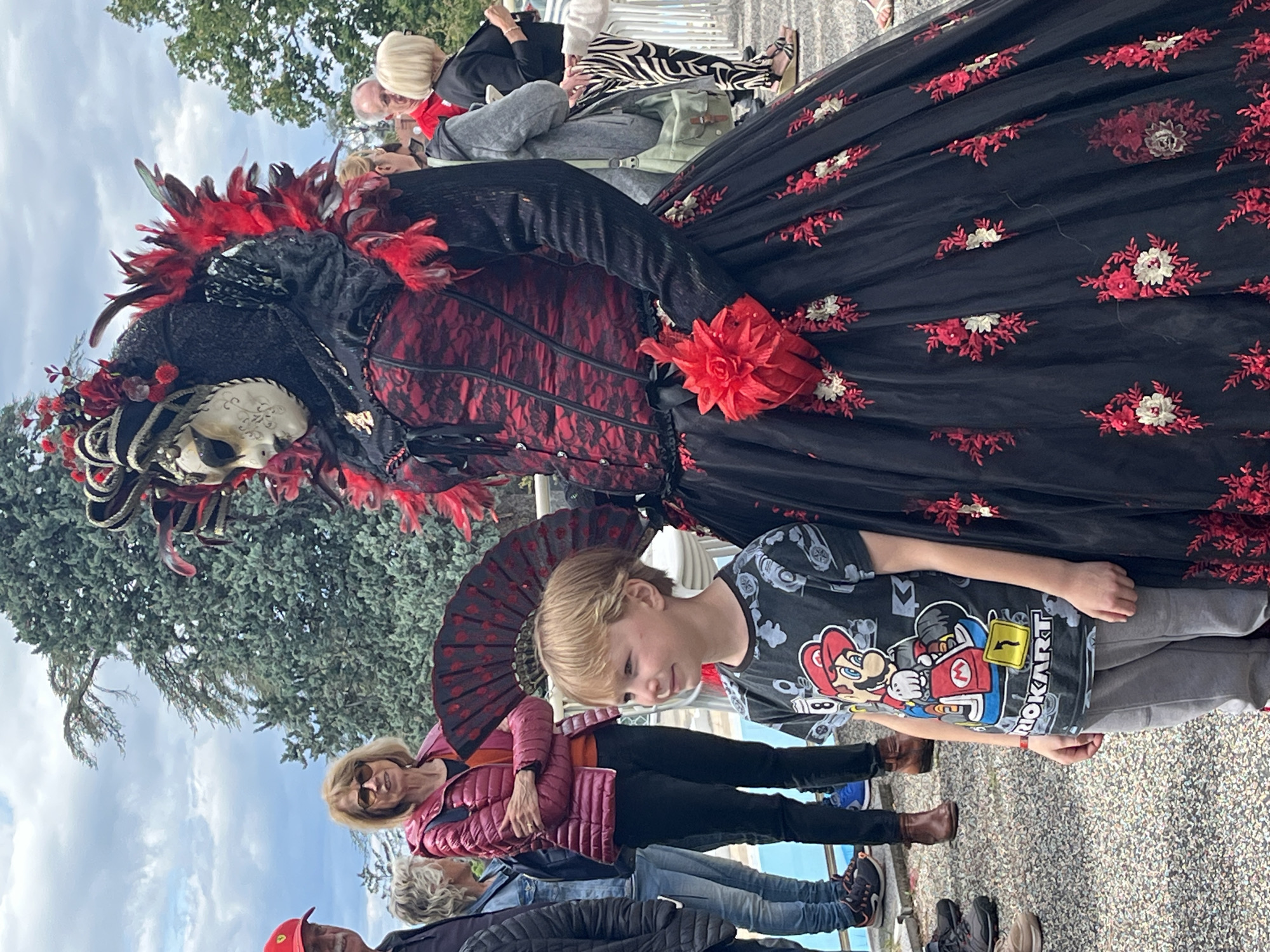 A medium-shot photo shows a person in elaborate carnival attire standing with a young boy. The person is wearing a black and red gown with floral embroidery and a feathered headdress, along with a white Venetian mask. The boy is wearing a grey Mario Kart T-shirt and gray pants. The background shows spectators of the carnival event, including a woman in a red jacket holding a black and red fan. The setting appears to be a public space with trees and a pebbled ground surface in Saint-Galmier, France.