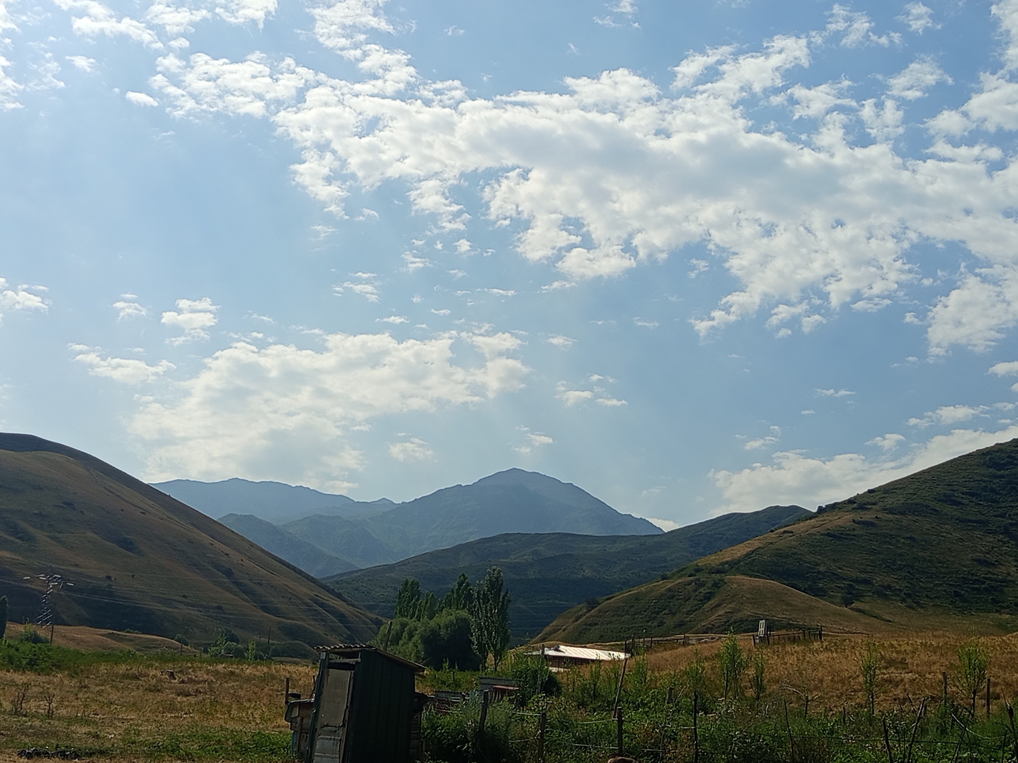 The image shows a landscape in Jardy-Su, Kyrgyzstan, featuring rolling hills and mountains under a partly cloudy sky. In the foreground, dry grass covers the ground, and a small, dilapidated shed-like structure is visible on the left. There is also a wooden fence with some greenery. Several tall trees stand in the middle ground, with a small building with a red roof behind them. The background is dominated by layers of mountains, with the furthest peaks appearing hazy and blue. The overall scene depicts a rural, possibly agricultural, environment. There are no people or animals visible in the image.