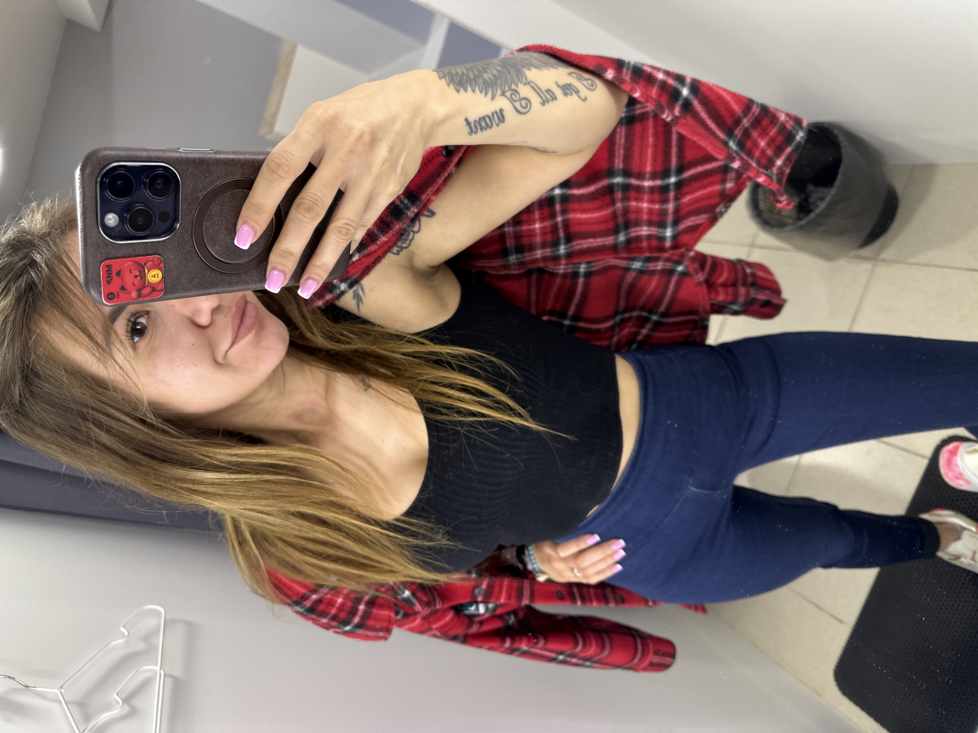 A woman with long brown hair is taking a selfie in a dimly lit fitting room. She is wearing a black crop top, dark blue leggings, and a red and black plaid shirt draped over her shoulders. Her right hand is holding a brown iPhone, and her left hand is on her hip. She has tattoos on her right arm and shoulder. The fitting room has a gray wall, a white hanger, and a black trash can. There is a black mat on the floor. The floor is tiled. The woman's toenails are painted pink. The woman has fair skin and a light smattering of freckles.