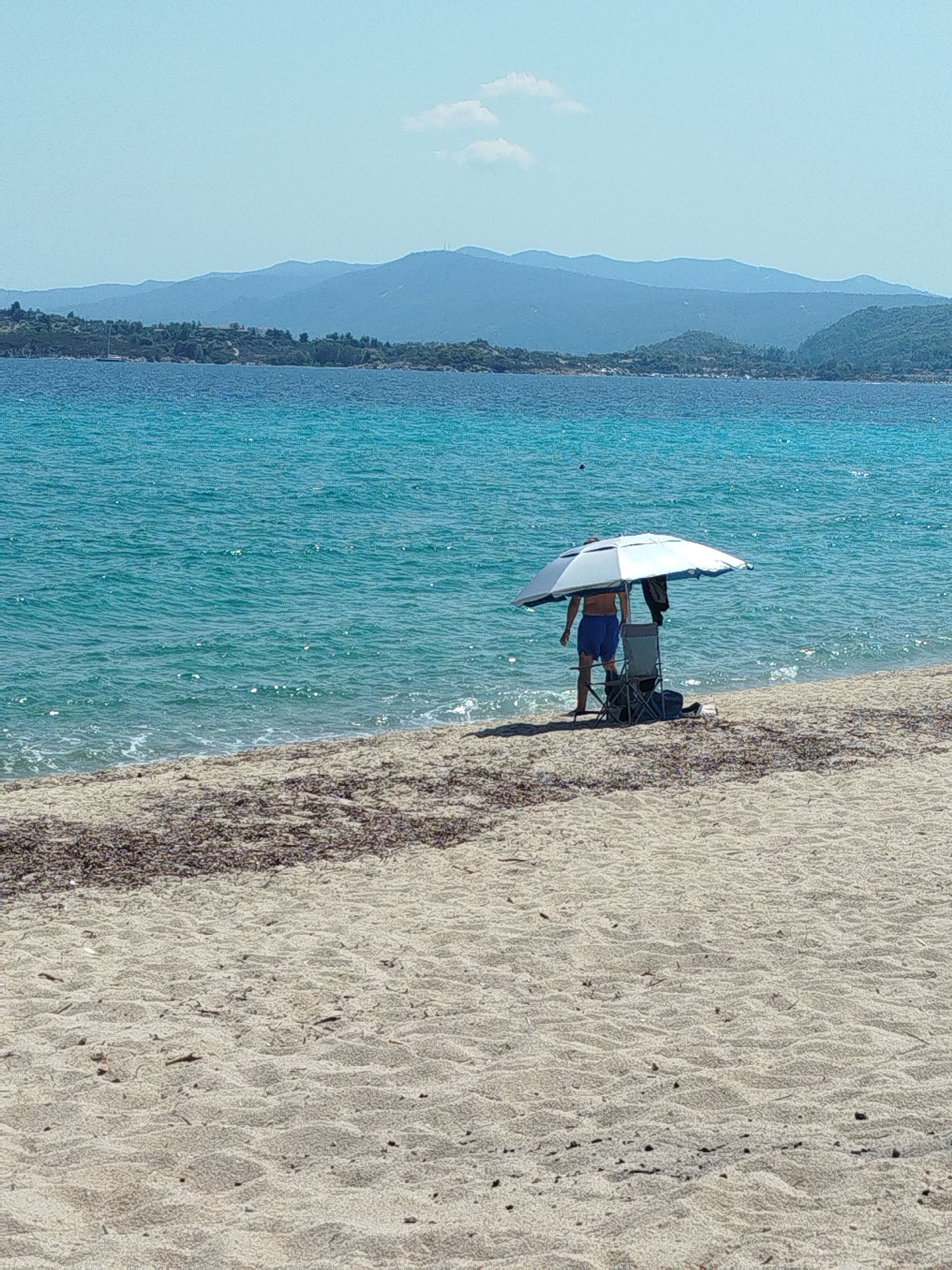 The image depicts a serene beach scene in Sithonia Municipal Unit, Greece, under a clear blue sky. In the foreground, a sandy beach stretches towards the turquoise waters of the Aegean Sea. A person stands under a large, open beach umbrella, partially obscured from view. The individual is wearing blue shorts and appears to be facing the sea. Next to them, a foldable beach chair is set up. Scattered on the sand closer to the water are strands of dark seaweed. In the background, a lush green coastline with trees and hills is visible, and beyond that, a series of blue mountain ranges fade into the horizon. The sea is calm with gentle waves lapping the shore.
