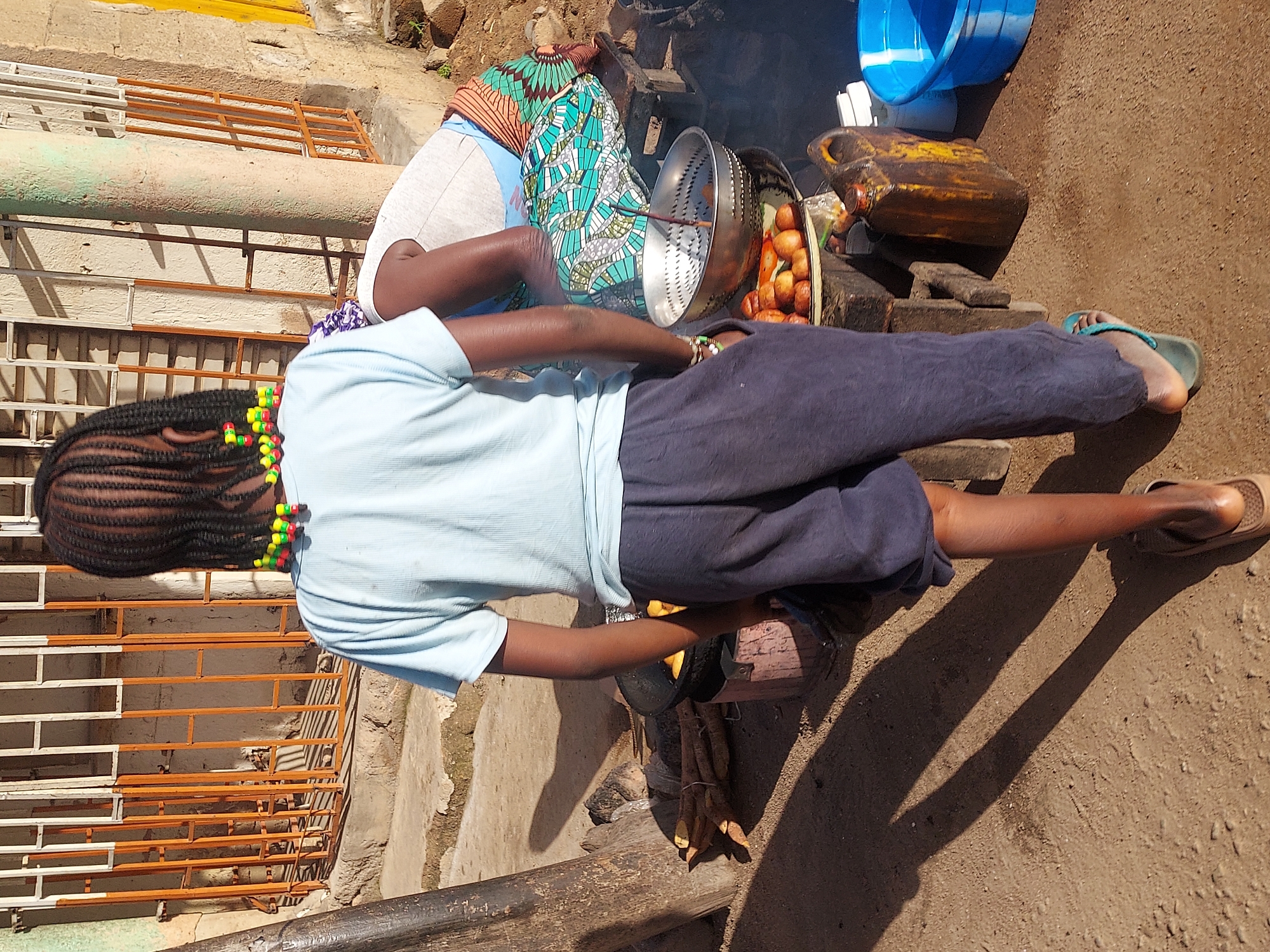This bright, sunny outdoor scene in Jos, Nigeria, captures a food preparation activity, likely at an informal street stall. In the foreground, a person with their back to the viewer wears a light blue t-shirt and dark blue trousers. Their braided hair is adorned with distinctive red, yellow, and green beads. They are positioned near a cooking setup.
Behind this individual, a second person is partially visible, seated on the ground and dressed in a vibrant patterned wrapper over light-colored clothing. This person appears to be tending to a fire where food is being cooked. Central to the activity is a large metal strainer holding a quantity of freshly fried items, possibly local snacks like puff-puff or fried yams, next to a larger pile of the same food. A dark yellow jerrycan, often used for cooking oil, and a bright blue plastic basin are also visible.
The ground is bare earth, and long, distinct shadows cast by the bright sunlight indicate it is daytime, likely mid-morning or early afternoon. In the background, there are light-colored walls and metal grates or fences painted in white and orange. Some raw root vegetables, possibly cassava or yam, are tied together and lie near the cooking area. One person wears teal flip-flops, and the other has light brown sandals. No legible text is visible within the image. The scene offers a glimpse into daily life and local food preparation practices in a Nigerian street setting.