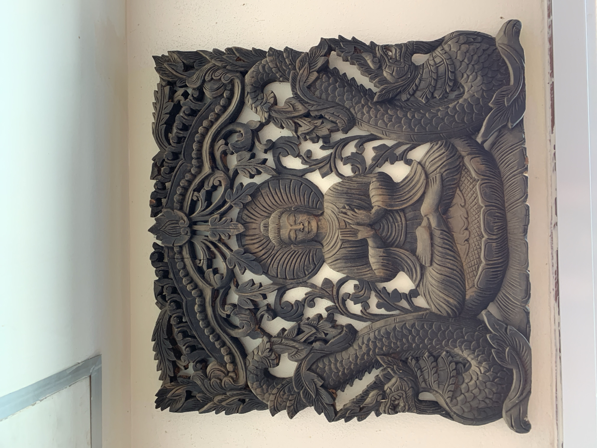 A carved wooden panel depicting a central figure surrounded by dragons adorns a wall in Baan Tai, Thailand. The panel is rectangular and features intricate details with the dragons at the top and bottom of the central figure. The panel is mounted on a pale wall.