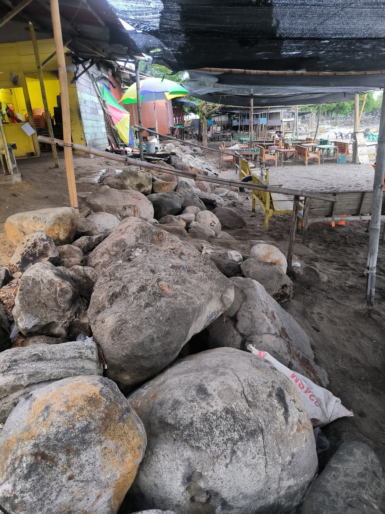 This detailed image captures an outdoor, informal beachside or riverside establishment in Lhokseumawe, Indonesia. The foreground is occupied by a substantial accumulation of large, irregularly shaped boulders, primarily grey and brown with some exhibiting yellowish or orange coloration and dark, damp patches. A white woven sack with red lettering, including "24CM," is visible nestled among these rocks. The ground throughout the visible area is composed of dark, coarse sand.
To the left, a bright yellow-painted wooden building, likely a shop or service area, is partially visible, with stacked plastic chairs discernible inside. Adjacent to this structure, a multi-colored beach umbrella (red, green, yellow, blue, white stripes) is partially furled, and another smaller, yellow and green umbrella stands nearby. Various bamboo poles and rough wooden planks serve as structural supports for the surrounding constructions.
The central and right portions of the scene feature an open-air seating area covered by a large, dark mesh or netting canopy, suspended by a network of bamboo and wooden poles. Underneath this canopy, numerous rustic wooden tables and chairs, some painted in yellow or light blue, are arranged on the sandy ground. At least two individuals are observed seated at these tables. In the background, additional similar structures extend along the shore, with scattered green vegetation and a distant view of what appears to be water or a further expanse of the coastline under an overcast sky.