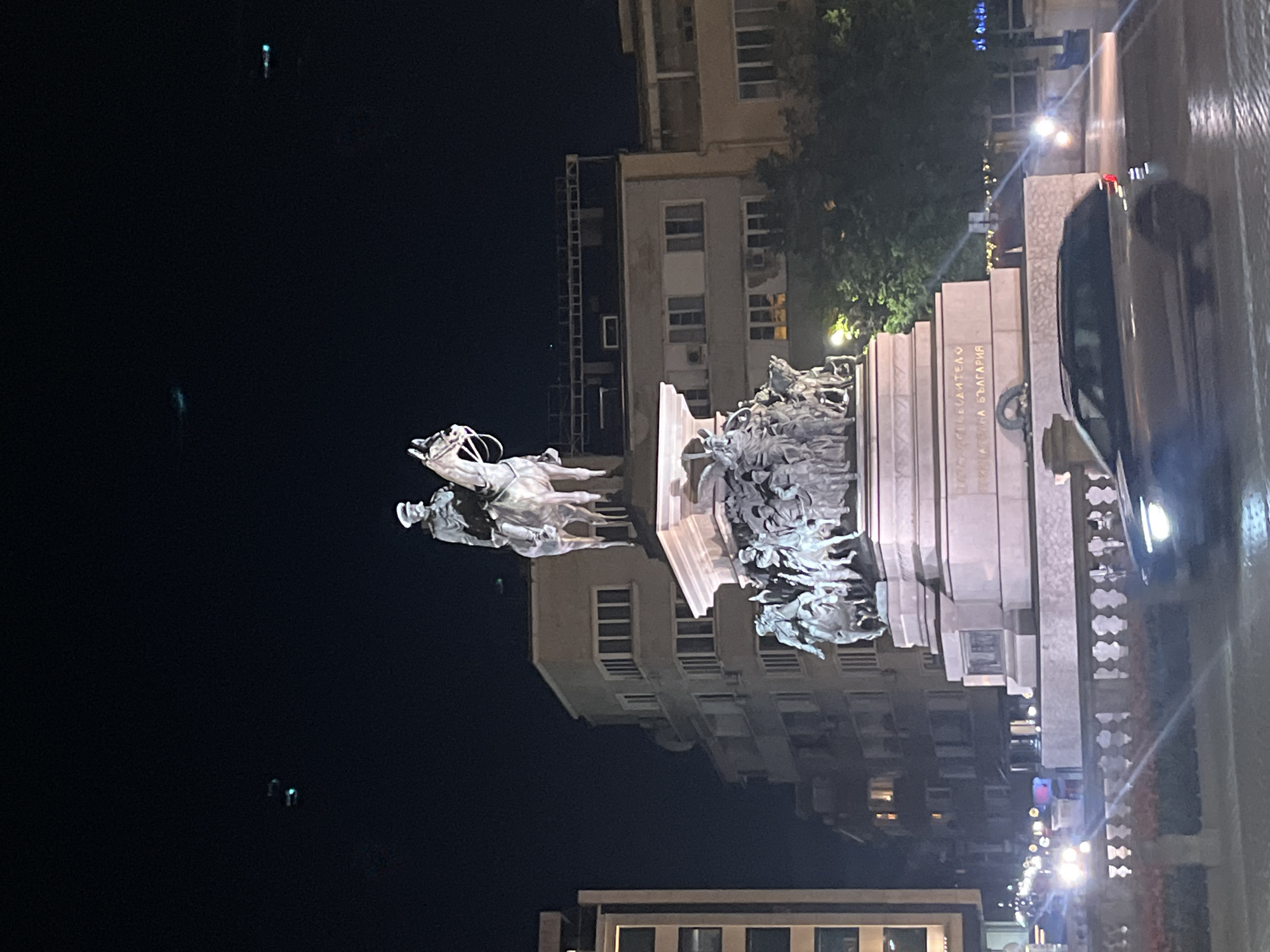 The image displays a prominent equestrian monument in Sofia, Bulgaria, captured at night. The central subject is a grand bronze equestrian statue of a rider on a rearing horse, positioned atop a substantial, multi-tiered stone pedestal. The base of the monument is adorned with intricate sculptural groups featuring numerous human figures, some appearing as soldiers and others as civilians, depicted in various dynamic poses.
Visible on the monument's base is Cyrillic text, reading "НА ОСВОБОДИТЕЛИТЕ / НА БЪЛГАРИЯ," which translates to "TO THE LIBERATORS OF BULGARIA," identifying it as the Monument to the Tsar Liberator.
The scene is set at night, characterized by a dark sky and artificial illumination from streetlights and lights emanating from nearby buildings. In the background, several multi-story urban buildings are visible, their windows glowing in places. Some trees and foliage are also present. To the right of the monument, a dark-colored car is partially visible, indicating a street or road adjacent to the monument. The overall atmosphere is calm, with the monument serving as a focal point under the night sky.