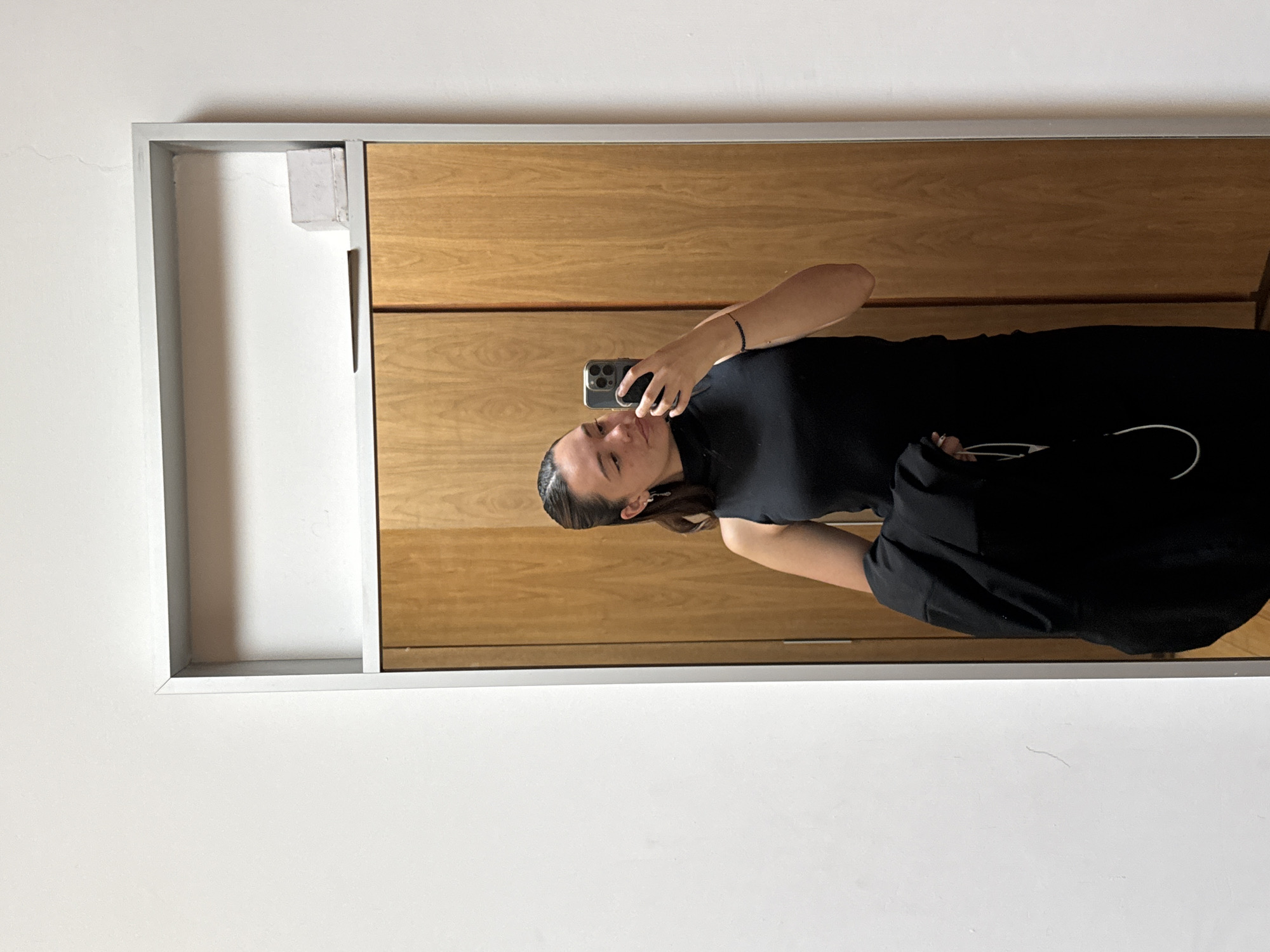 An indoor mirror selfie captures a young adult woman in San Bernardino Tlaxcalancingo, Mexico. The woman is reflected from the waist up, wearing a form-fitting black sleeveless or short-sleeved top with a high neckline, and possibly black pants or a skirt, part of which is visible. Her dark hair is styled back. She holds a dark smartphone, likely an iPhone due to the visible camera module, in her right hand, aiming it towards the mirror to take the photo. A black beaded bracelet adorns her right wrist. Her left hand rests casually on her hip. She looks towards the phone with a subtle, pleasant expression or slight smile.
The setting appears to be a room with a white wall. The mirror is framed in grey, and to its right, a wooden surface, possibly part of a cabinet or closet, is visible. The lighting is bright and even, suggesting daytime. A thin white cable or strap is faintly visible near her left side. There are no specific distinguishing features in the background to indicate the precise location within Mexico. No visible text or brand names are present in the image.