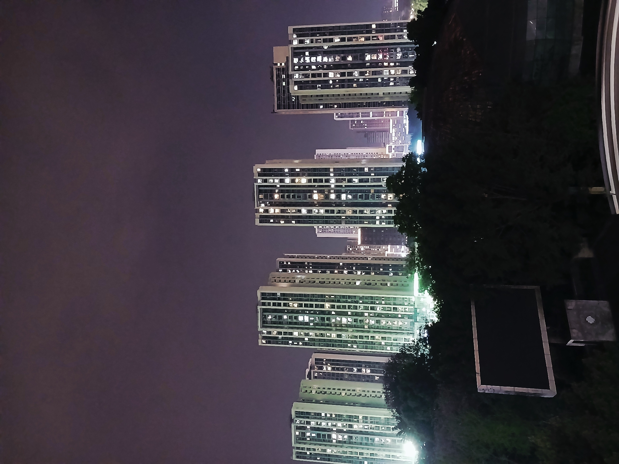 The image shows a nighttime view of high-rise apartment buildings in Chongqing High-tech Zone, China. The buildings are illuminated with lights in many windows. The sky is dark and overcast. In the foreground, there are trees and a structure (possibly a rooftop or building feature). A road or walkway with railings is visible on the right side.