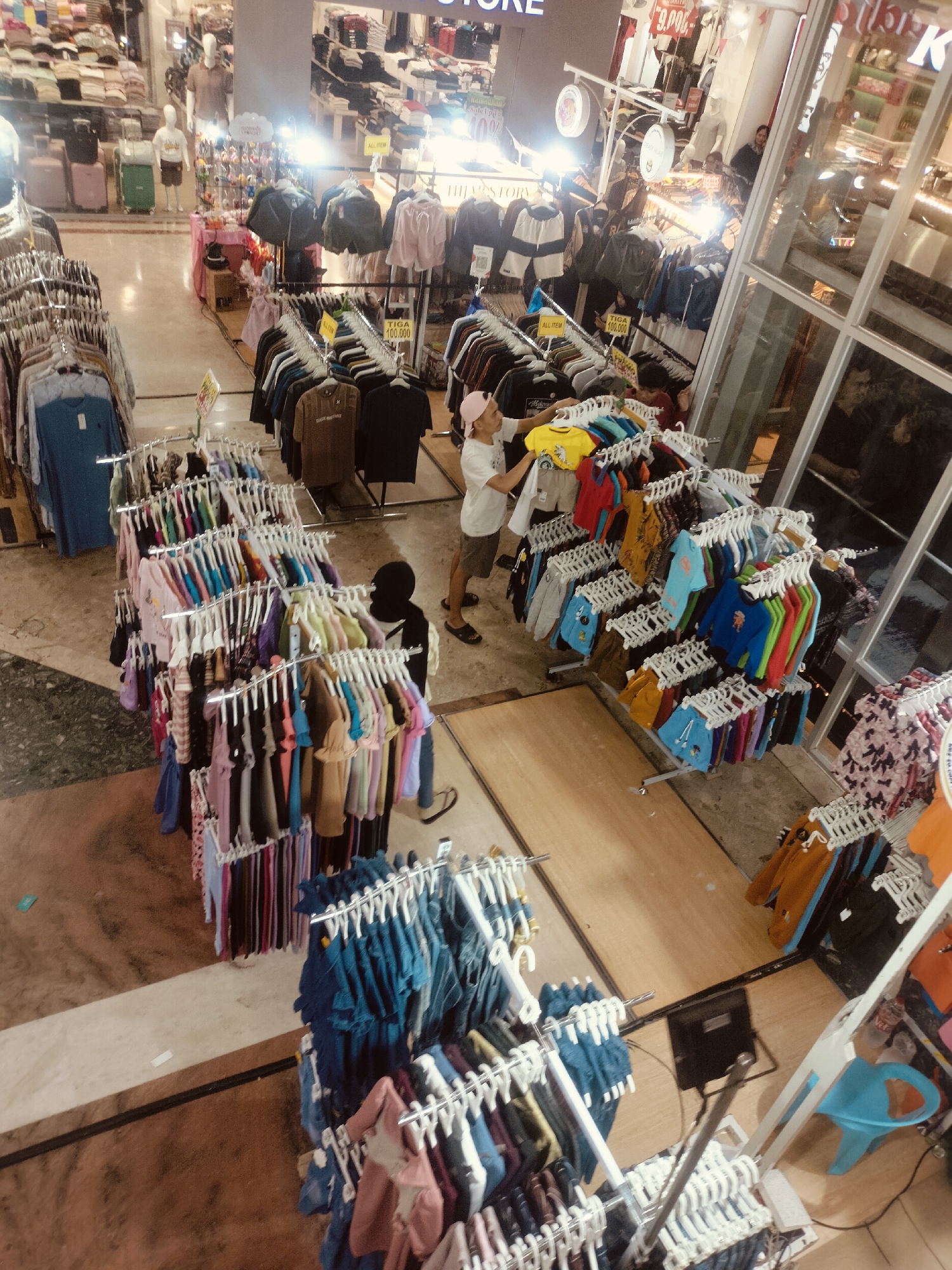 The image depicts a brightly lit indoor shopping mall in Surabaya, Indonesia, filled with clothing racks and displays. In the foreground, a young person wearing a pink cap and white t-shirt is browsing through a rack of colorful t-shirts. Beside them, another person is partially visible, wearing a black head covering and dark clothing. Various clothing items, including t-shirts, shorts, and jackets, are neatly arranged on metal racks, some with price tags attached. In the background, more clothing displays and mannequins are visible, as well as shoppers interacting and moving through the mall. The floor of the mall is a mix of tiled and wooden surfaces. The overall scene suggests a busy retail environment.