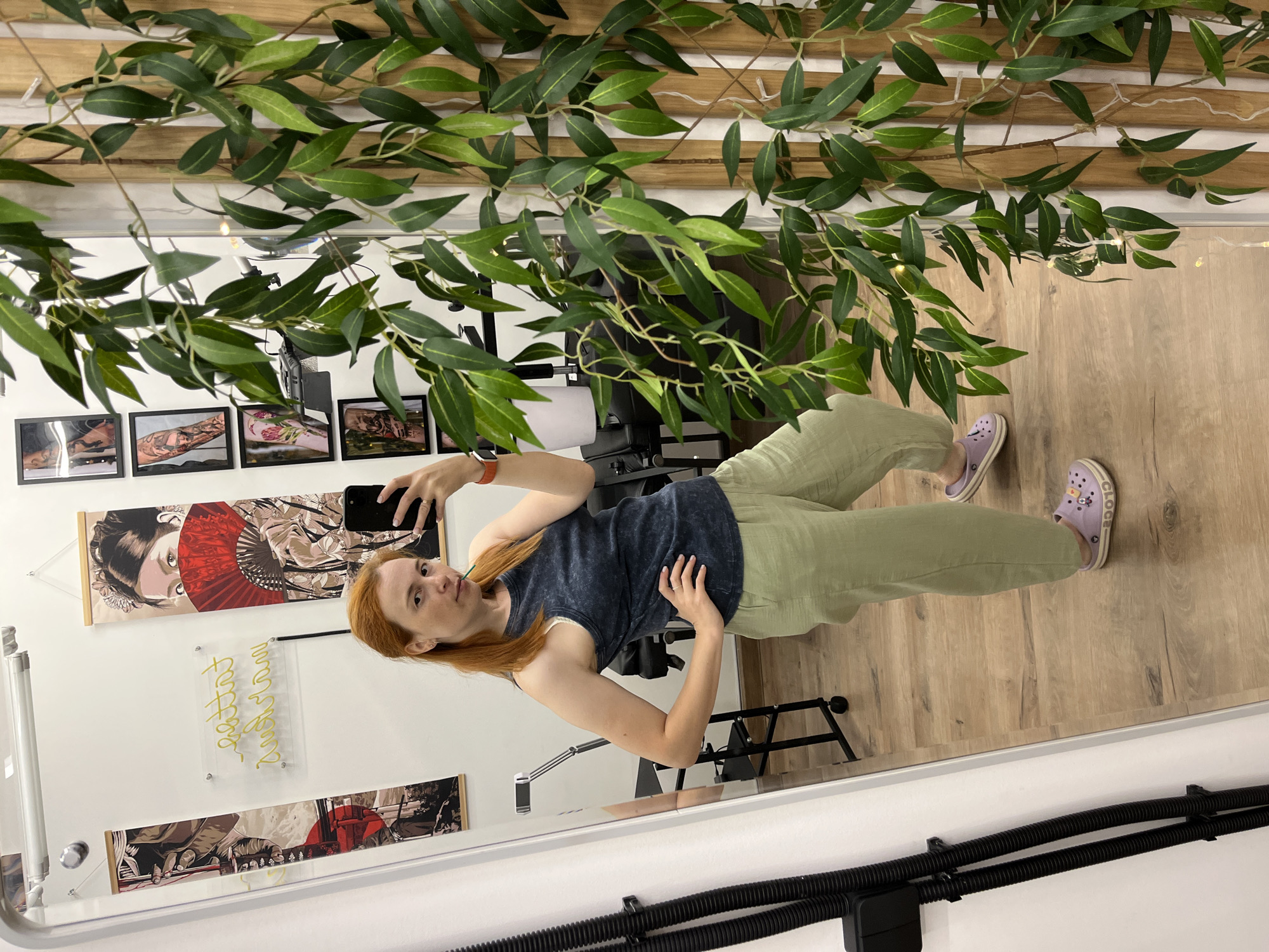 In a well-lit indoor space with wooden flooring and white walls, a woman with red hair is taking a mirror selfie. She is wearing a dark blue tank top and light green pants, accessorized with a red watch and purple clogs. In her right hand, she holds a smartphone, and a green object, possibly a straw, is in her mouth. Behind her, a tattoo parlor's decor is visible, including framed tattoo art on the wall and a neon sign that reads "Tattoo Land" in cursive. A large mirror reflects the scene, and a tall artificial plant with green leaves partially obscures the right side of the image. There are no indications of the specific location in Chaikovsky, Russia.