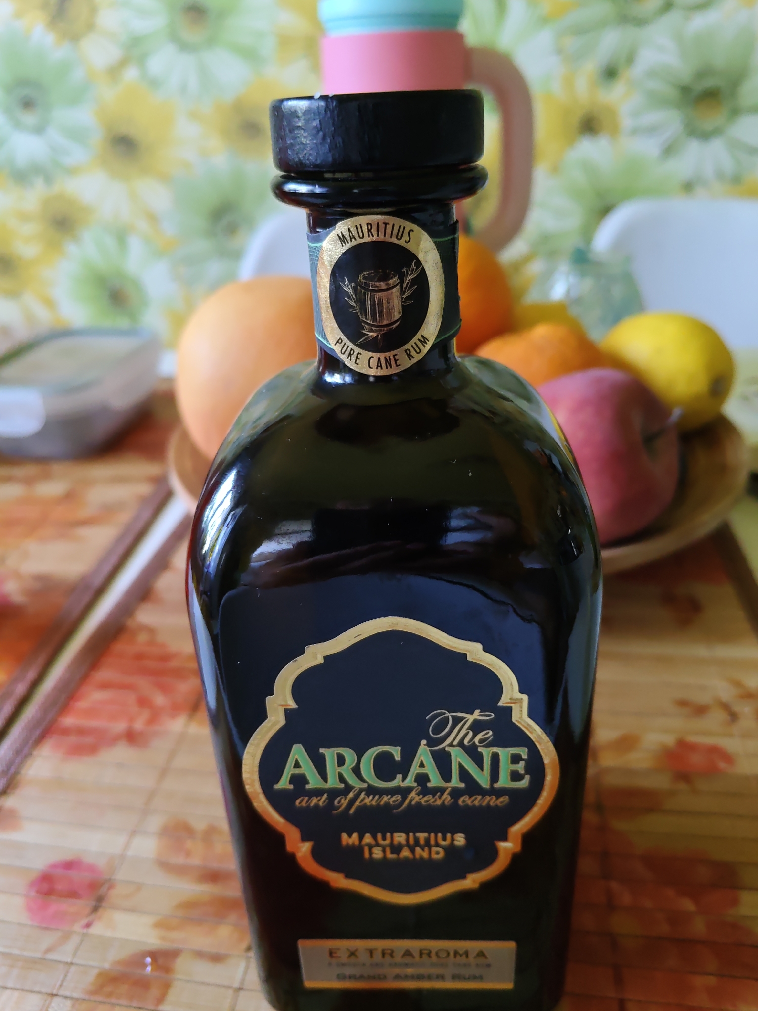 The image shows a close-up of a dark glass bottle of rum, labeled "The Arcane" and "Mauritius Island". The bottle has a black cap and a gold and dark blue label with ornate lettering. Behind the bottle, partially visible, are several fruits, including what appears to be an orange, a lemon, and a red apple, arranged in a bowl. The background features a wall with a floral pattern in shades of yellow and green. The scene is set on a table with a bamboo placemat. There is no indication of the city of Rostov-on-Don, Russia, in the image or its content.