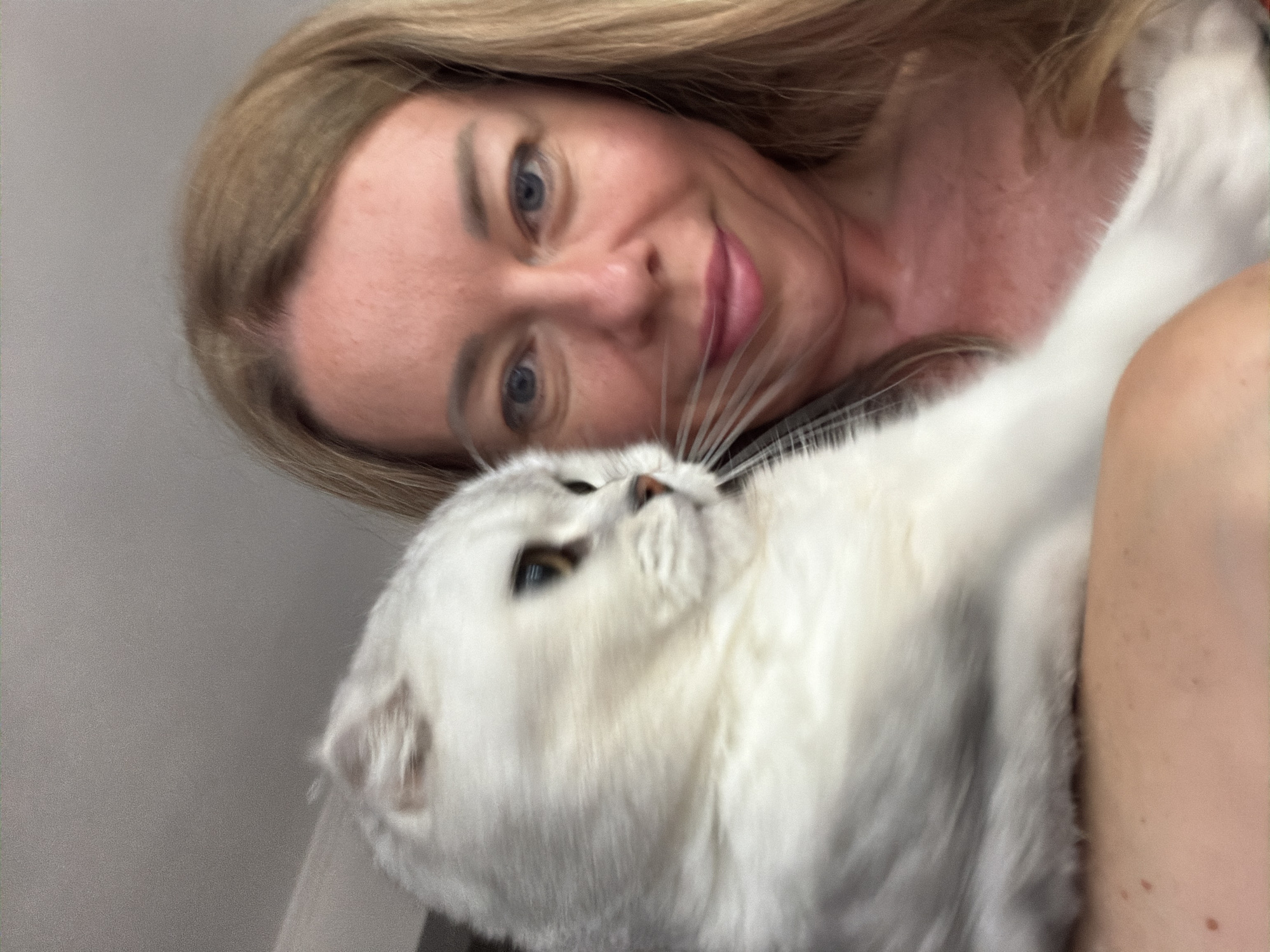 A woman with long blonde hair and blue eyes is holding a white cat. The cat is on the left side of the image, facing right, and its fur is blurred due to motion. The woman is on the right side of the image, looking at the camera and smiling. Her skin is lightly tanned and she is wearing a light-colored top. The background is a plain, light-colored surface. There is no discernible information to suggest the location is Rome, Italy.