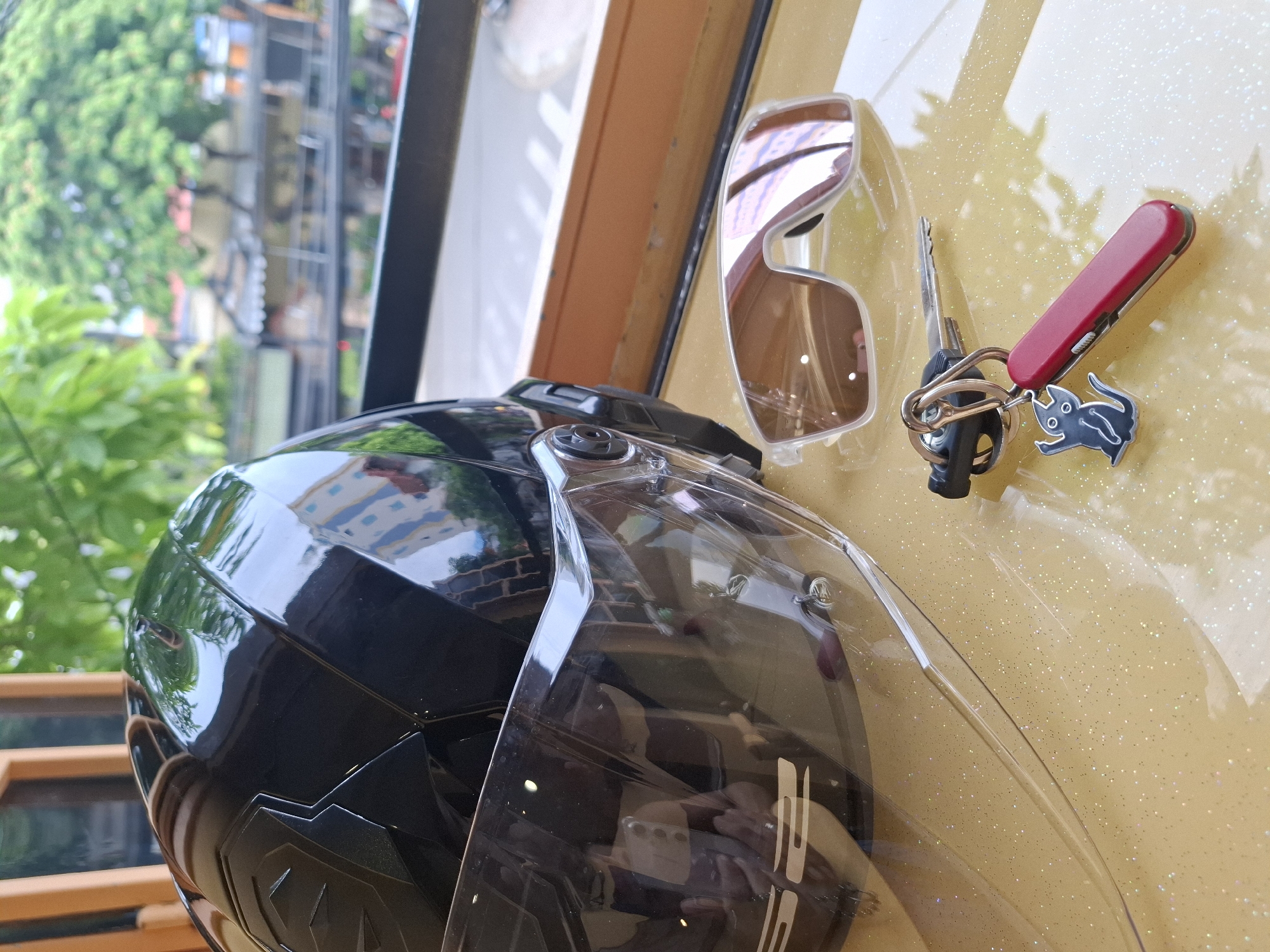 The image depicts a still life of motorcycle accessories placed on a yellow, glittery surface. The objects include a black motorcycle helmet with a clear visor, a pair of light-colored sunglasses with reflective lenses, and a set of keys attached to a keychain featuring a red multi-tool and a black charm shaped like a cartoon cat. The background is blurred, showing a view from an elevated position, possibly a balcony or window, looking out onto a street scene in Nha Trang, Vietnam. Elements visible in the background include buildings, trees, and a road with faint traffic. The lighting suggests an outdoor, daytime setting.