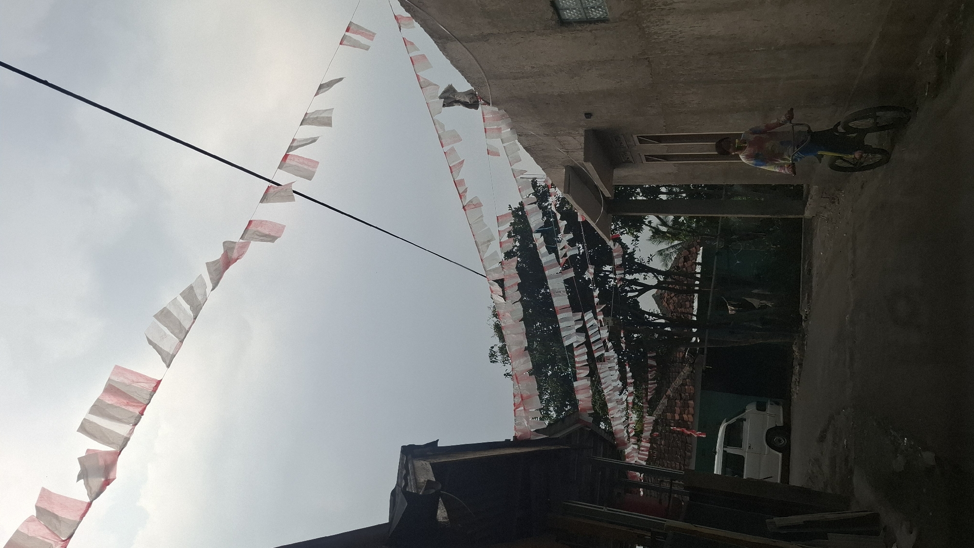 The image shows a street scene in Sukasari, Indonesia. Red and white Indonesian flags are strung across the street, with more flags draped over the roofs of houses. A person is riding a bicycle down the street, and a white van is parked on the side of the road. The sky is overcast. The architecture suggests a residential area with modest buildings.