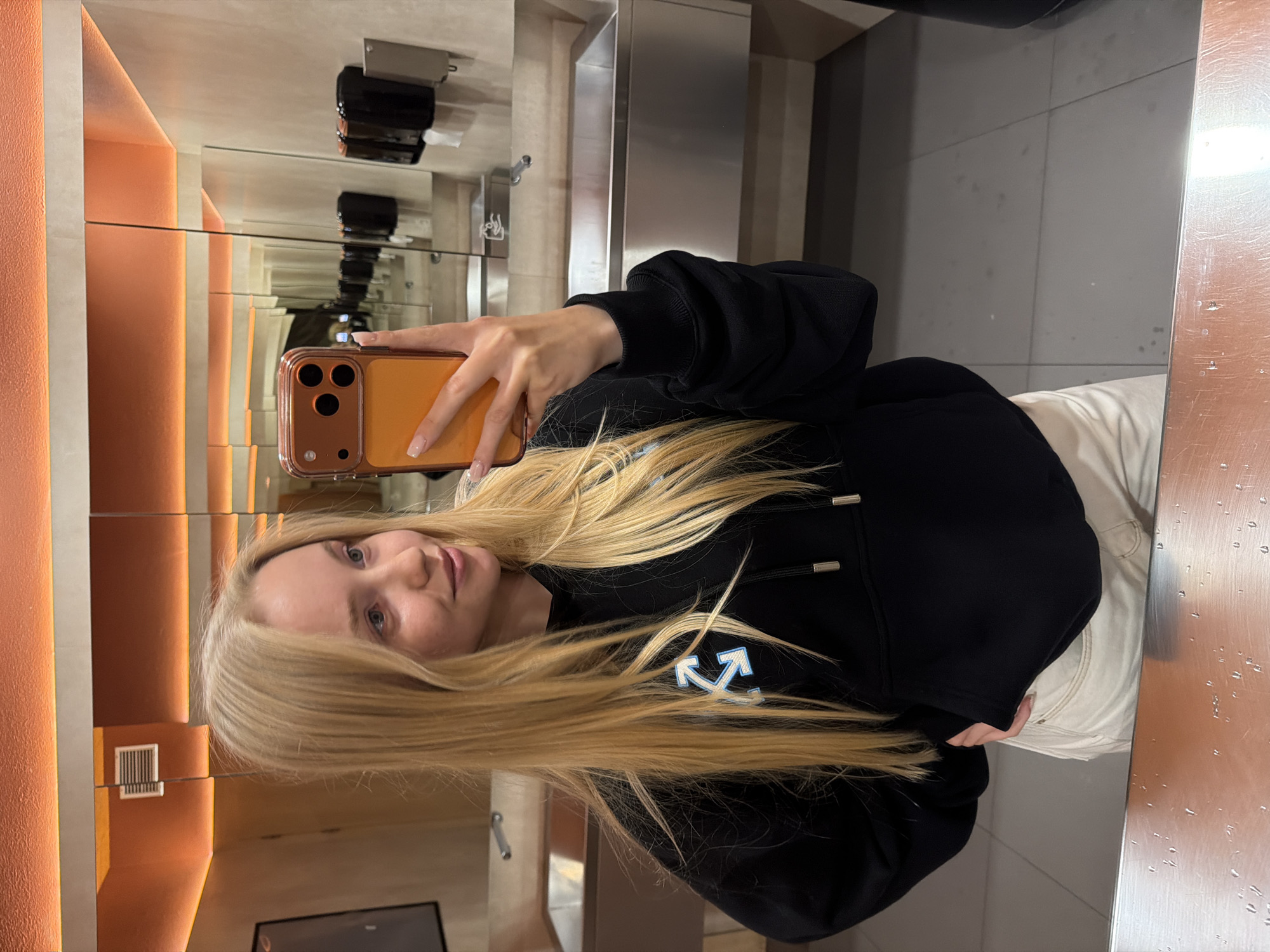A woman is taking a selfie in a public restroom. She is holding a phone with a brown case up to the mirror, which reflects the scene behind her in an endless loop. The woman has long blonde hair, fair skin, and is wearing a black hooded sweatshirt with a light blue and white logo on the front and white pants. The restroom has a modern design with an orange accent wall, stainless steel fixtures, and gray floor tiles. Soap dispensers are visible on the wall. The restroom appears to be clean and well-maintained.