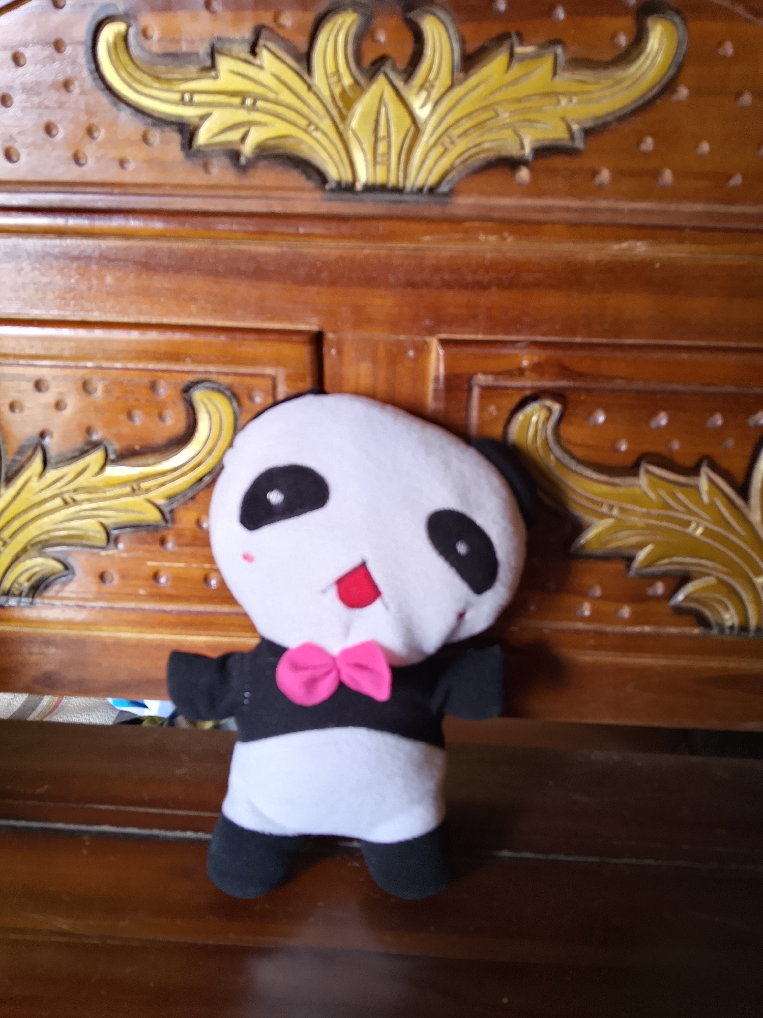 A plush panda toy is positioned in front of a wooden cabinet with ornate golden carvings. The panda is white with black eye patches, ears, and feet, and is wearing a black vest with a pink bow tie. Its mouth is open, revealing a red tongue. The background cabinet has a dark wood finish and features intricate, leaf-like golden embellishments. The scene appears to be indoors.
