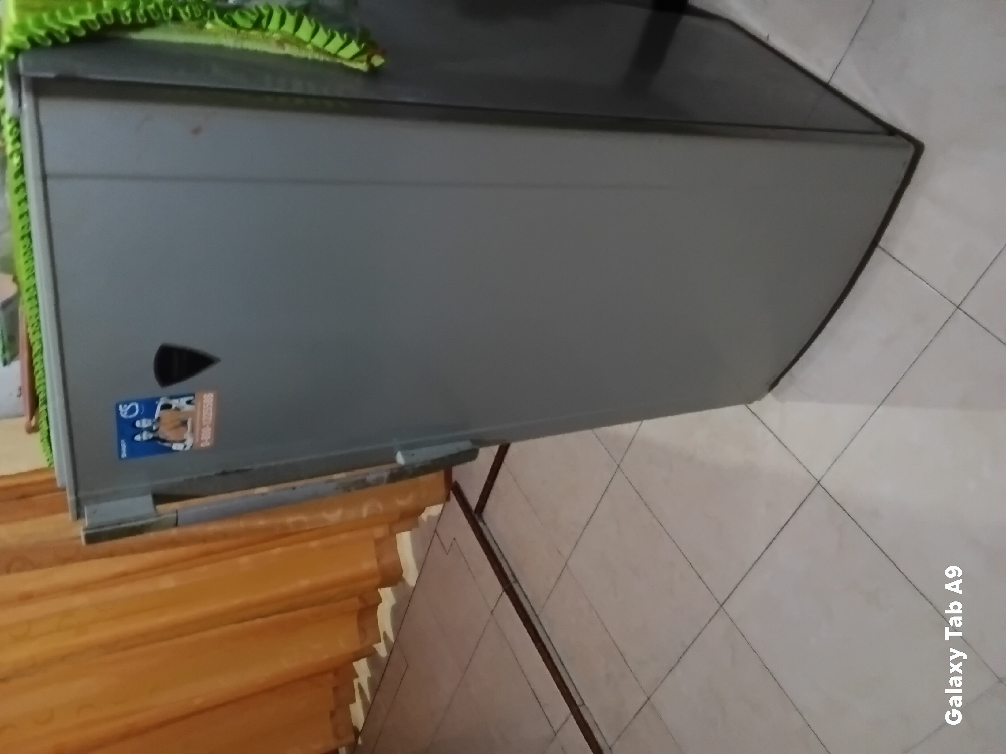 A light gray refrigerator stands in a room. It has a sticker on its door featuring two individuals in athletic wear and a phone number. A green cloth with a patterned trim hangs over the top of the refrigerator. To the left of the refrigerator, orange curtains cover a window. The floor is tiled with light-colored square tiles. The image is branded with "Galaxy Tab A9" in the lower left corner.