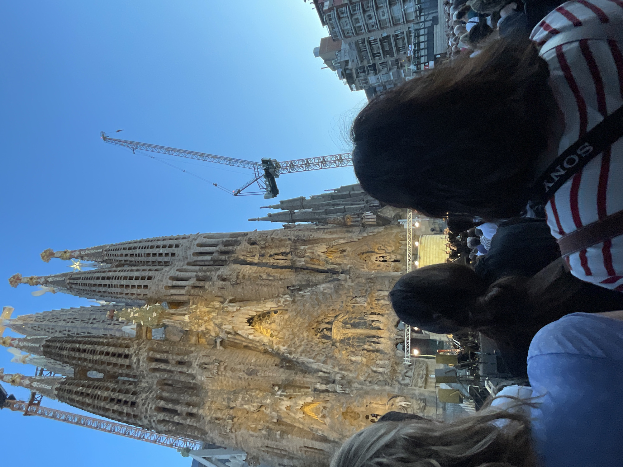 The image depicts the Sagrada Familia in Barcelona, Spain, during an evening event, likely a concert. The basilica's facade is illuminated, showcasing its intricate Gothic and Art Nouveau architecture. A large construction crane is visible to the left of the basilica, indicating ongoing work. In the foreground, the backs of several people are visible, suggesting they are spectators attending the event. Some of these individuals are wearing casual clothing, and one person has a camera strap with the "SONY" logo across their shoulder. In the middle ground, a stage with musicians and equipment is set up in front of the basilica. The background consists of apartment buildings and a clear blue sky. The overall scene suggests a cultural event taking place in a prominent urban setting.