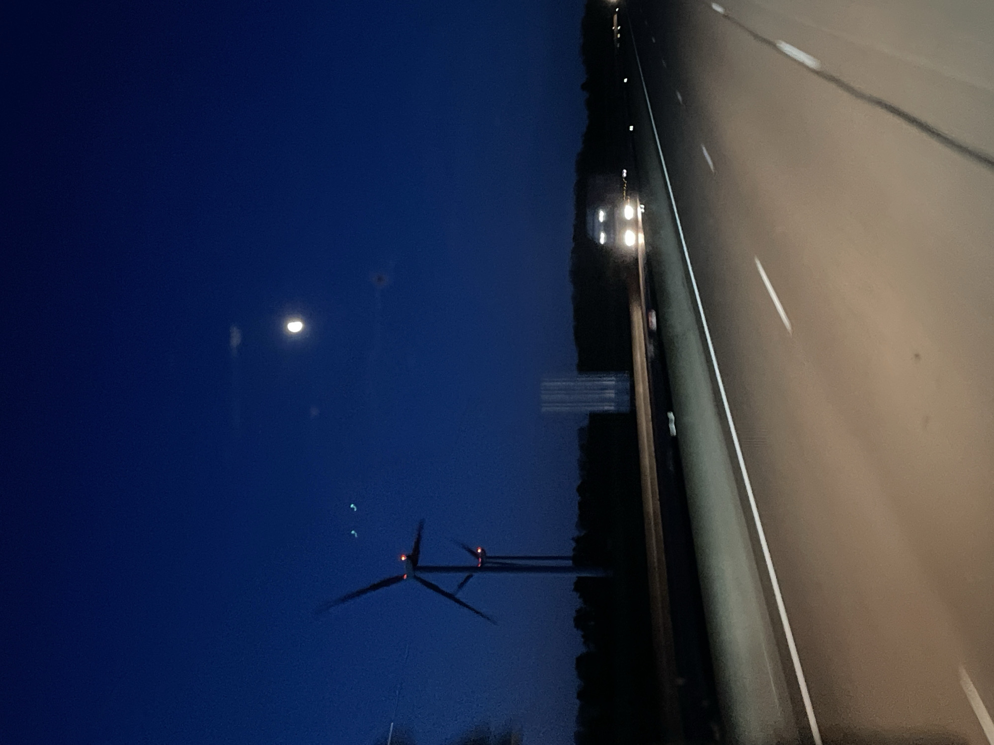 The image shows a view from inside a vehicle, likely a car, driving at night. To the right is a multi-lane highway with white lane markings. The shoulder of the road is lined with trees and utility poles. Across the road is a field with wind turbines. The sky is dark blue, with a bright full moon visible.
Given that this is Vesvres-sous-Chalancey, France, this might be the A31 autoroute.