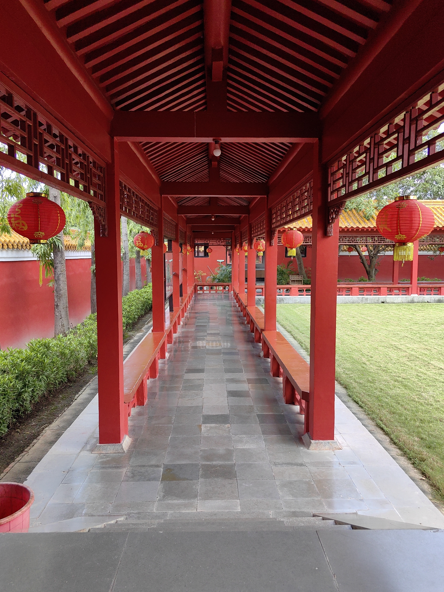 The image captures a long, red-painted covered walkway with a tiled floor, leading into the distance. Red lanterns hang from the ceiling and posts, some featuring yellow lettering. The walkway is flanked by wooden benches on one side and a red wall with lush green bushes and trees on the other. To the right of the walkway, a well-maintained green lawn stretches out. The architecture suggests a traditional East Asian or South Asian style, possibly a temple or a heritage site. The location is identified as being in Lumbini Bazaar, Nepal.