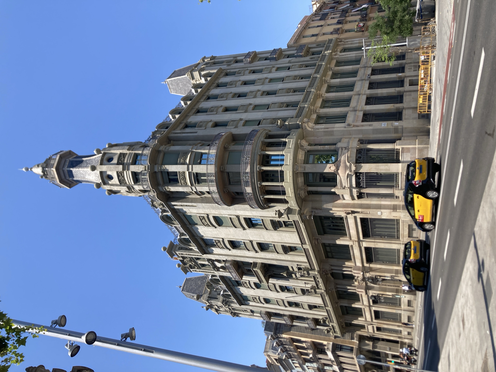 The image depicts a street view in Barcelona, Spain, featuring an ornate, historic building with a distinctive turret and balconies. The building's facade is constructed of stone with numerous windows and decorative elements. In the foreground, two yellow and black taxis are visible on a paved street. A lamppost with multiple lights stands on the left side of the image, and trees are present along the sidewalk. The sky is clear and blue, indicating a sunny day. The scene suggests an urban environment with architectural heritage.