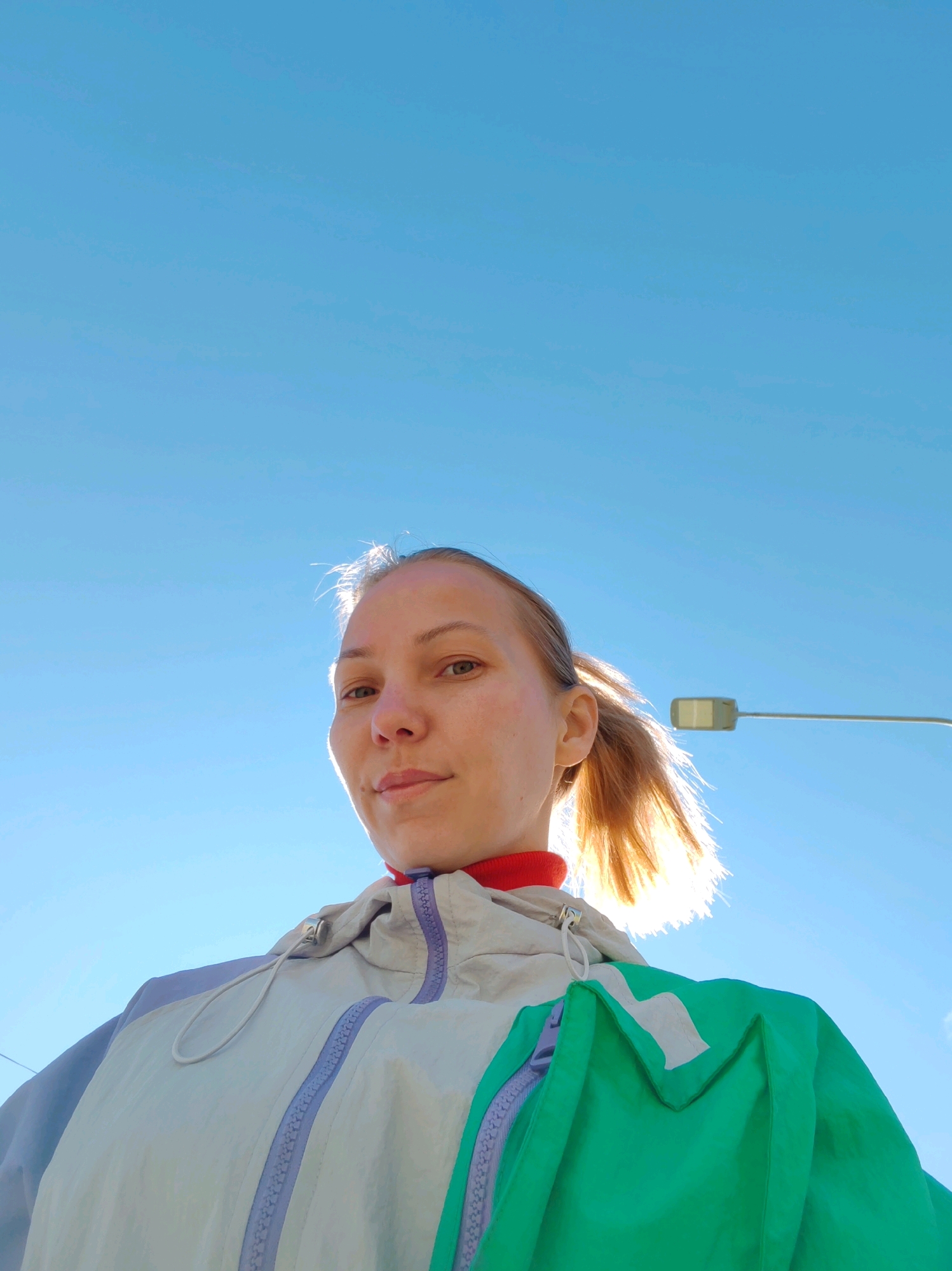 The original image shows a low-angle, chest-up shot of a young woman with blonde hair pulled back in a ponytail, wearing a multi-colored jacket and a red collar. She is outdoors, with a clear blue sky as the background. A street light is visible in the upper right portion of the image. The lighting suggests it is daytime and sunny, with the sun positioned behind the woman's head, creating a halo effect around her hair. The woman is looking directly at the camera with a neutral expression. The image was taken in the city of Chukhlem, Russia.