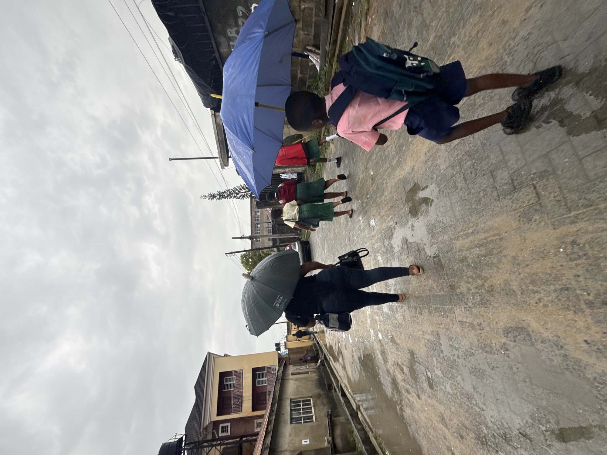 A street scene in Ado, Nigeria, features several individuals walking in the rain. A young boy with a blue umbrella wears a pink shirt and blue shorts, carrying a green backpack. Behind him, a group of children in school uniforms walk together. A woman with a dark-colored umbrella wears dark clothing and sandals. The street is wet, reflecting the overcast sky. Buildings line the street, including a two-story building with windows. Power lines are visible overhead.