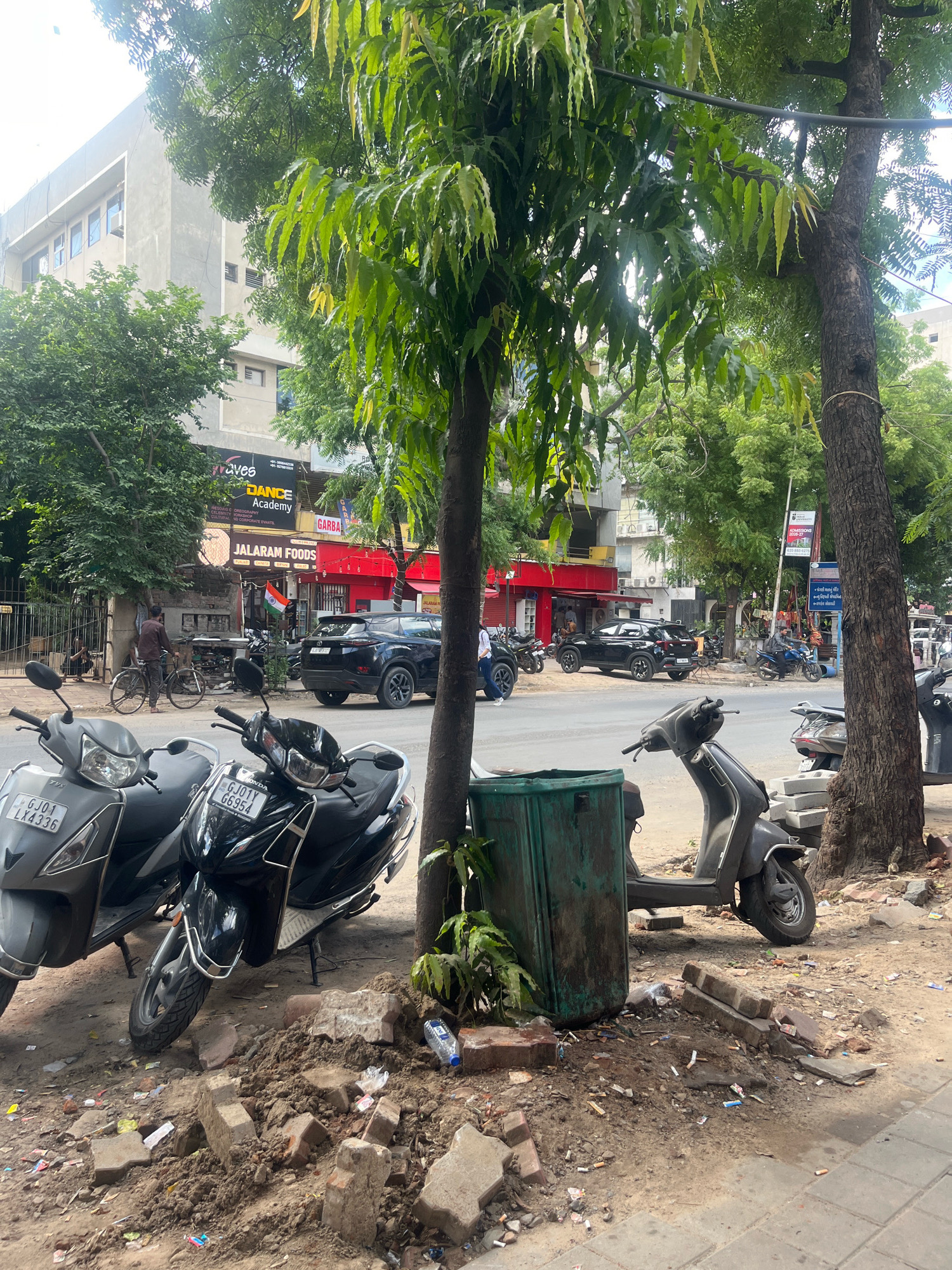 The image depicts a street scene in Navrangpura, India. In the foreground, two scooters are parked on the side of a road. The scooter on the left is grey, and its license plate reads "GJ01 LX4336". The scooter on the right is black, and its license plate reads "GJ01Y GG954". A green trash bin is situated to the right of the black scooter, and a pile of bricks and debris is located between the two scooters.
The background features a street with vehicular traffic, including a black SUV. Several shops are visible along the street, with signage including "JALARAM FOODS" and "DANCE ACADEMY". Trees line the sidewalk, and a building with multiple windows is present behind the shops. Several people are visible, including a man with a bicycle near a shop entrance and others walking on the sidewalk. The overall environment appears to be an urban commercial area on a cloudy day.