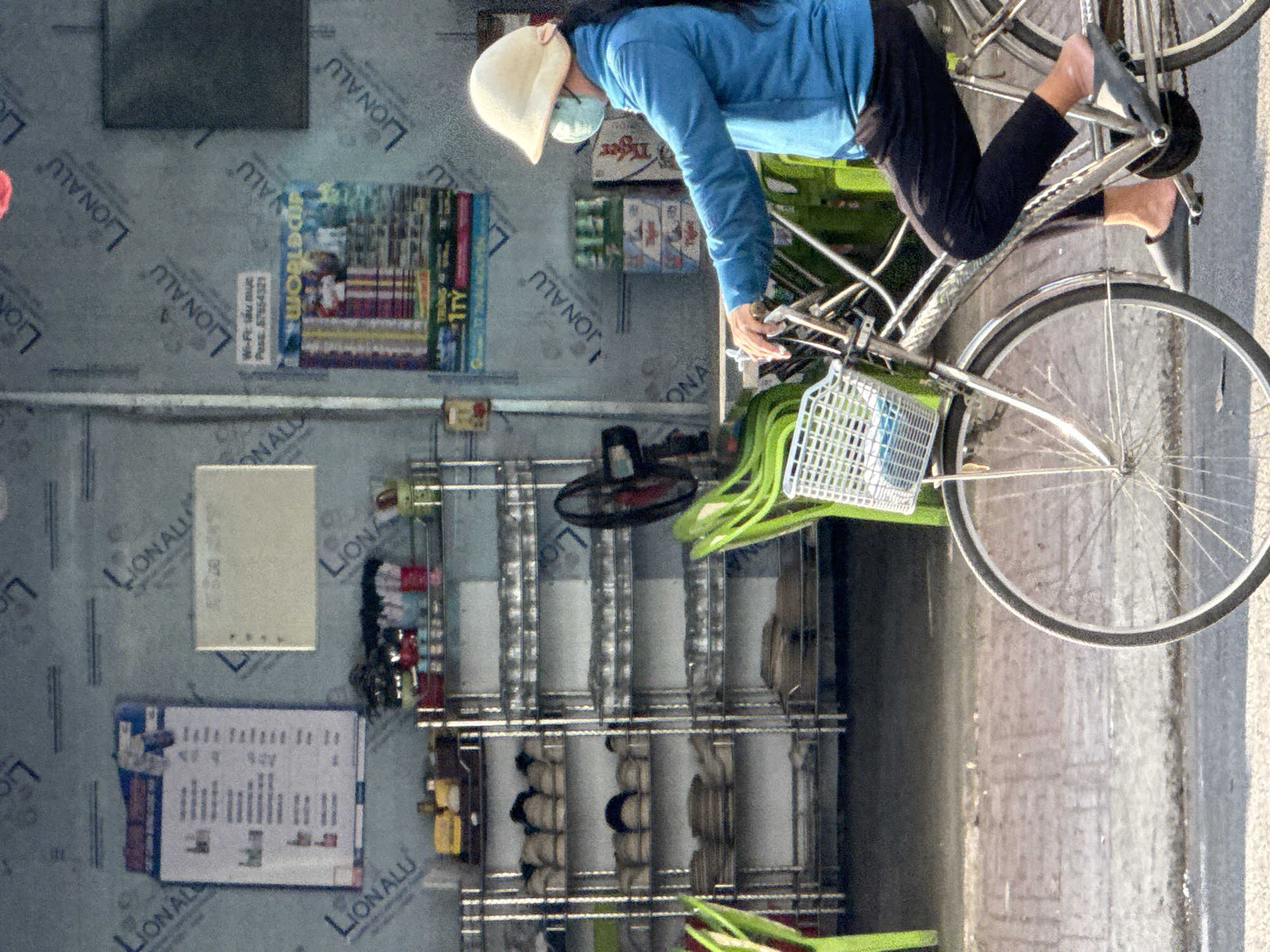 A person wearing a blue shirt, black pants, and a light-colored hat is riding a bicycle. The person is also wearing a mask. The bicycle has a white basket on the front. The scene appears to be outdoors, possibly in an alley or by a street, with a wall in the background covered in repeating text "LIONALL". There are shelves with various items on the wall behind the bicycle. Visible text on the wall includes "Wi-Fi tầu mục Pess: 57654321" and "WORLDONAU 1TY". The overall lighting suggests it could be daytime. The image is cropped and rotated. The background suggests an urban or commercial setting.