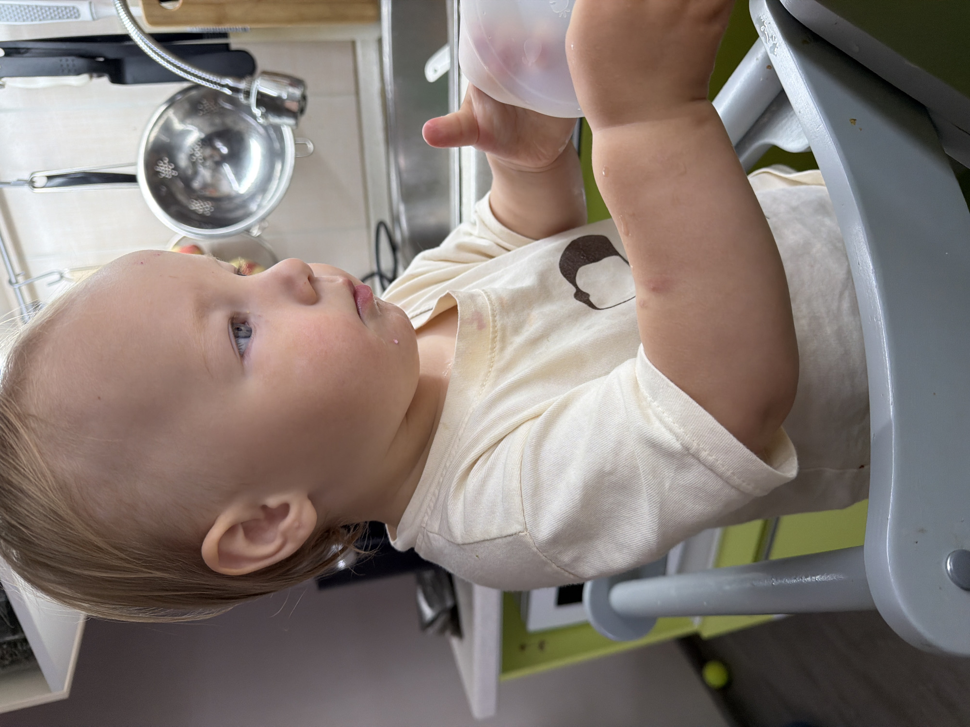The image shows a close-up, side profile of a young child, likely a toddler, sitting in a high chair. The child has fair hair and blue eyes and is wearing a light-colored t-shirt with a brown graphic. The child's mouth is slightly open, with a small pink mark visible on the lower lip, possibly from food or drink. The child's right hand is holding a clear plastic cup or lid, and there are water droplets on the child's arm and the high chair.
The background suggests a kitchen environment. A sink with a faucet is visible, along with various kitchen utensils hanging on a rack, including a metal colander. To the left of the sink, there is a green cabinet or counter. The overall lighting is bright, indicating an indoor setting during the day.
The child appears to be engaged in an activity involving the cup and possibly drinking or playing with water. The scene is clean and appears to be a domestic setting. The location of Chaikovsky, Russia, is not directly indicated by any visual elements in the image, but the description is provided as requested.