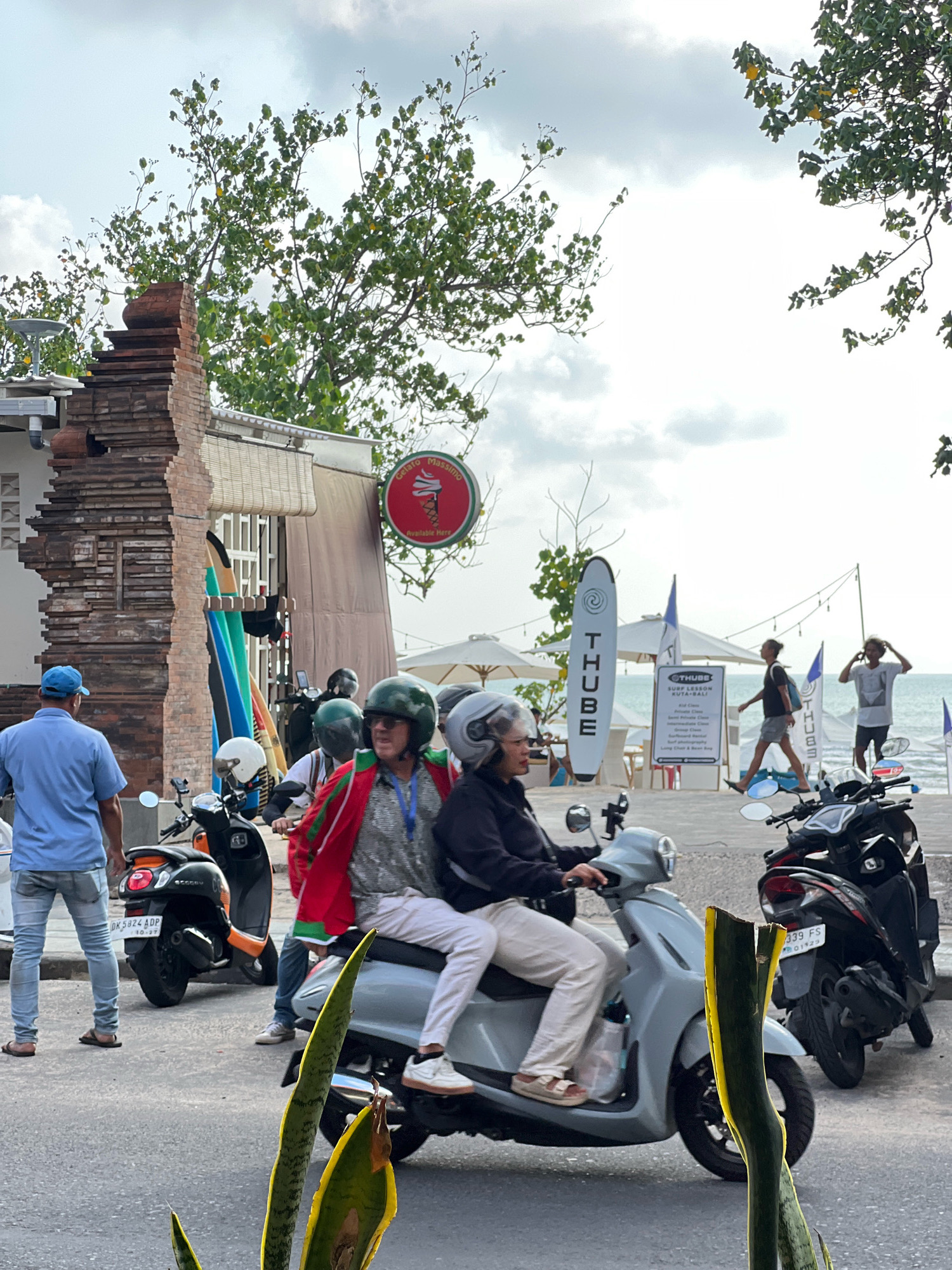 In Kuta, Indonesia, a street scene unfolds with a focus on a light gray scooter carrying two occupants. The front rider, a woman with a helmet, wears sunglasses and a dark jacket. Beside her, a passenger in light-colored pants and a helmet is seated. The scooter is in motion, moving towards the left of the frame. Behind this scooter, a black scooter is parked, its license plate reading "339 FS". Further left, a person wearing a blue shirt and blue jeans stands near an orange and black scooter, also parked. In the background, a row of surfboards stands upright against a building, and a red sign with an ice cream cone and "Gelato Massimo" is visible. Beyond the building, beach umbrellas, surfboards with the "THUBE" logo, and the ocean are present, indicating a coastal location. The sky is overcast.
