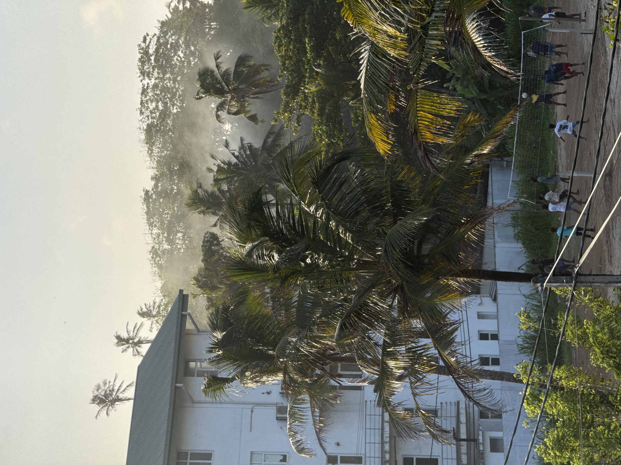 In Goviyapana, Sri Lanka, a group of young men are engaged in a volleyball game on a dirt court. The game is being played under a hazy sky, with the sun filtering through palm trees and a light mist or smoke in the background. A white building, possibly a dormitory or hotel, is visible on the left side of the frame, with several air conditioning units attached to its exterior. A volleyball net is set up in the center of the court, and players are spread out across it, actively participating in the game. Some players are reaching for the ball, while others are positioned defensively. The surrounding environment is lush and tropical, with dense vegetation and tall palm trees dominating the landscape. The overall scene captures a moment of everyday life and recreation in a tropical setting.