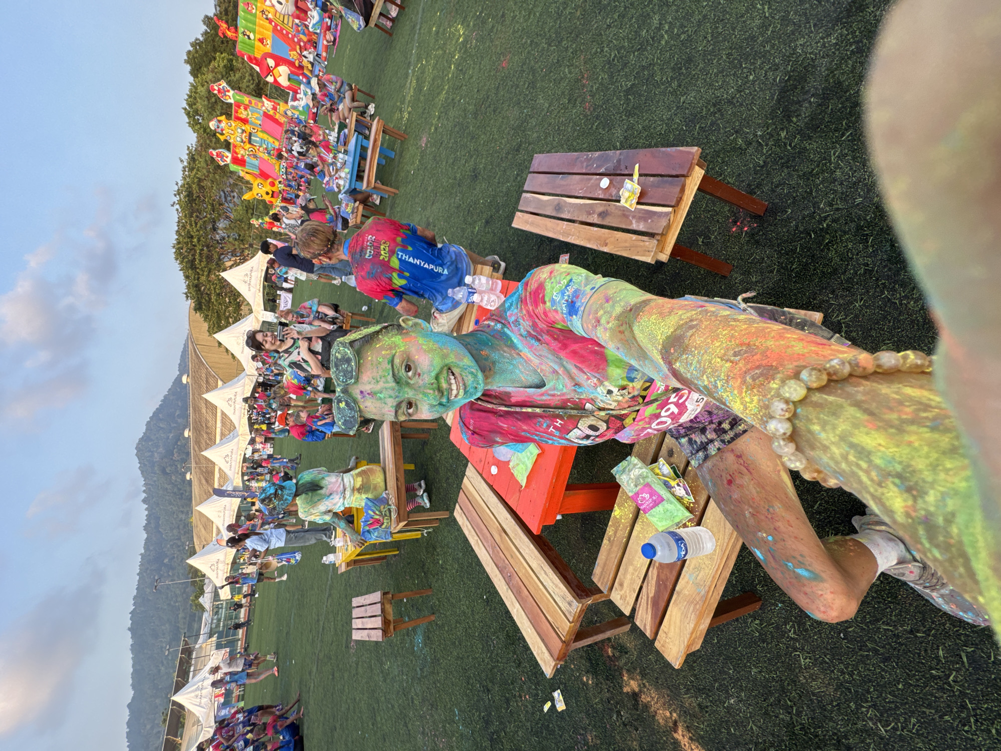 A woman sits on a picnic bench in a field in Thep Krasatti, Thailand. She is covered in colored powder and appears to be at a color run or festival. She is taking a selfie, her arm outstretched. In the background, there are rows of white tents and a crowd of people. There are some colorful inflatable structures in the distance.