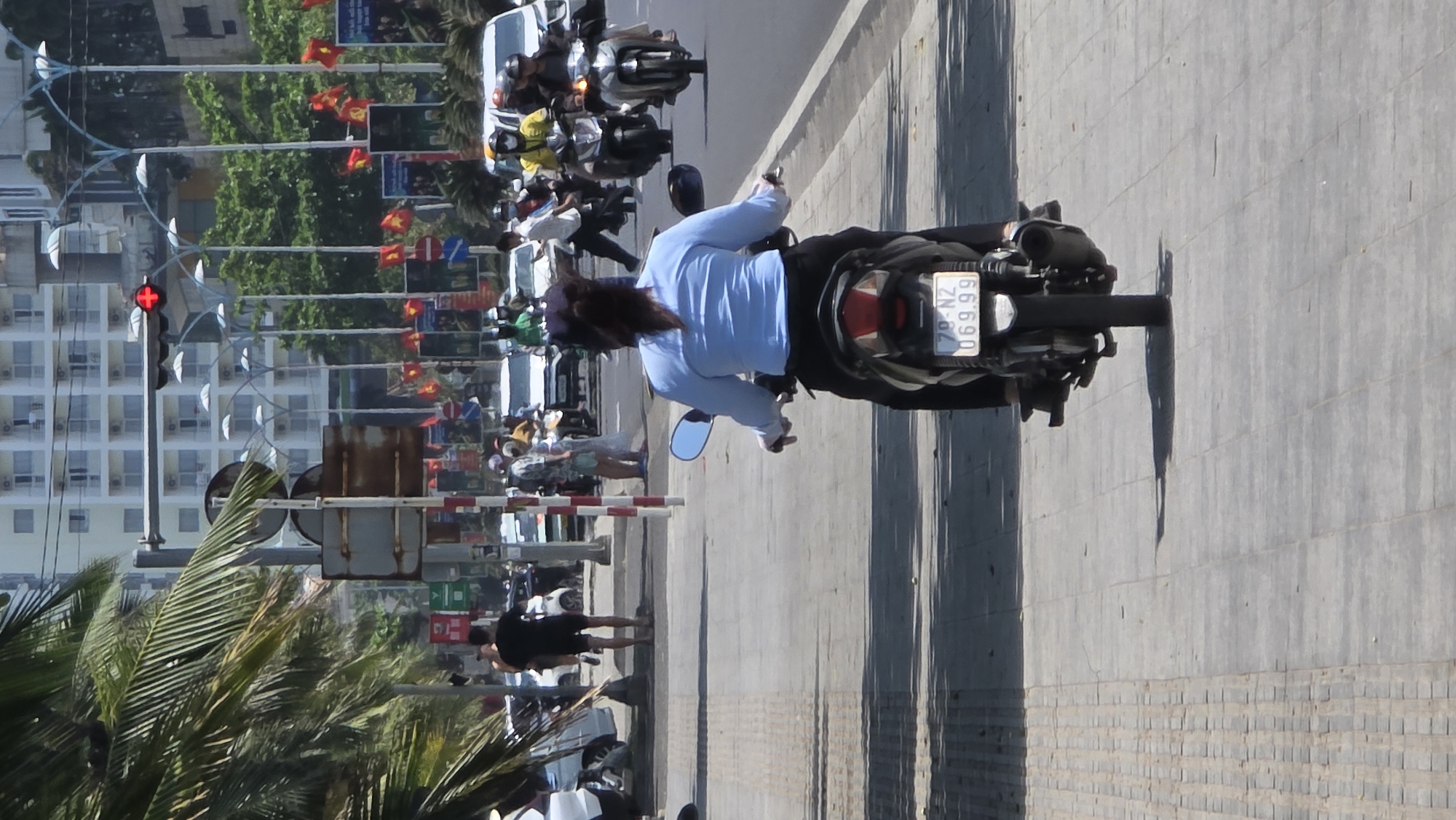 The image shows a street scene in Nha Trang, Vietnam. A person on a motorbike, facing right, is positioned on a sidewalk or paved area. The motorbike's license plate reads "79-N2 069.99." A row of red flags and traffic signs lines the street. A red traffic light is visible in the background, with a building facade behind it. Several pedestrians are visible on the sidewalk. A group of motorbikes are on the road. Palm trees are on the left.