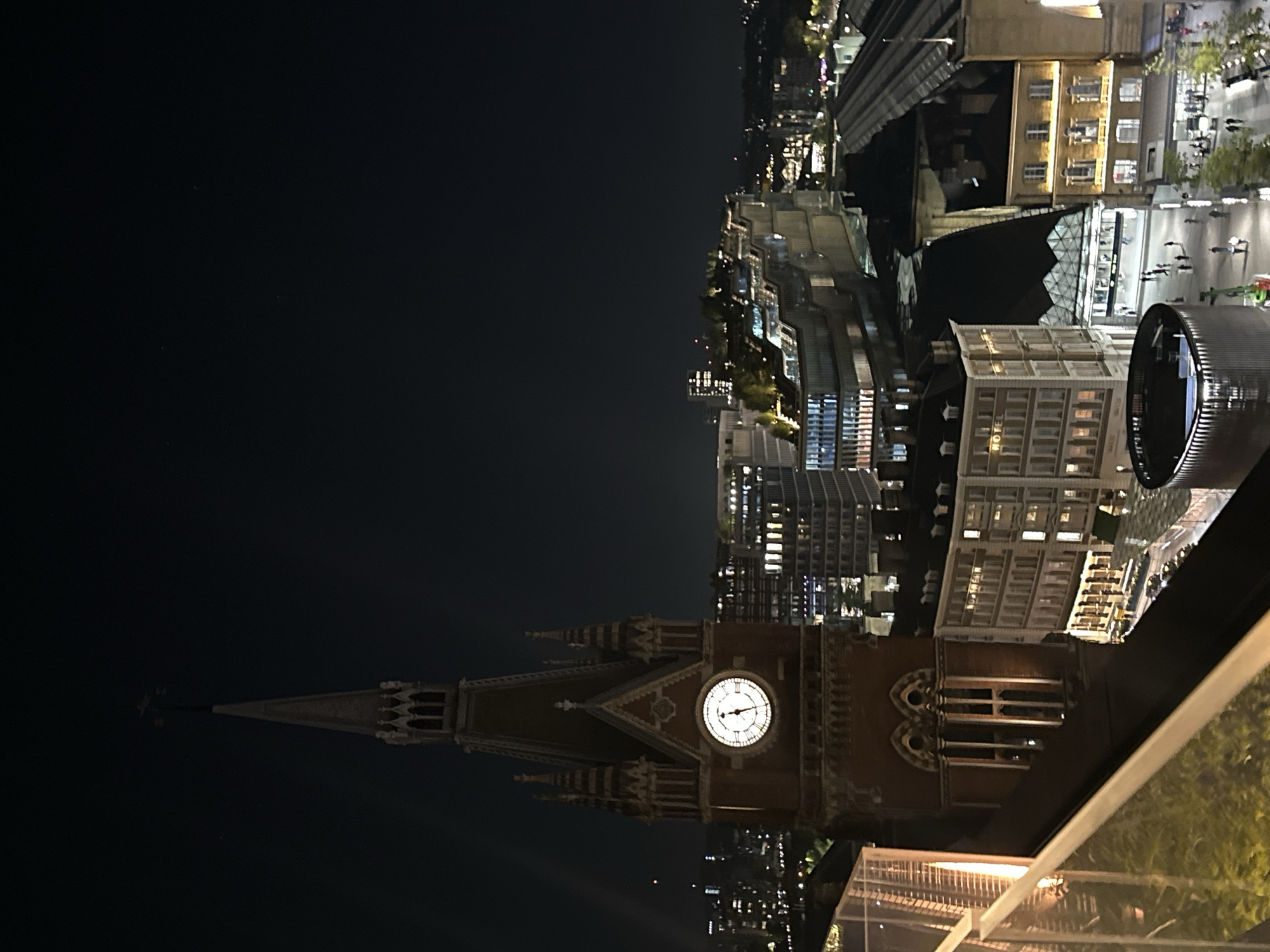 The image captures a nighttime view of the cityscape of London, United Kingdom. A prominent feature in the foreground is the St. Pancras clock tower, displaying its illuminated clock face. The time on the clock appears to be approximately 11:58. Beyond the tower stretches a modern urban landscape, dotted with illuminated buildings, including what appears to be a hotel. In the distance, pedestrians are visible walking along a well-lit street. The photograph is taken from an elevated position, possibly from a rooftop terrace, offering an overview of the area.