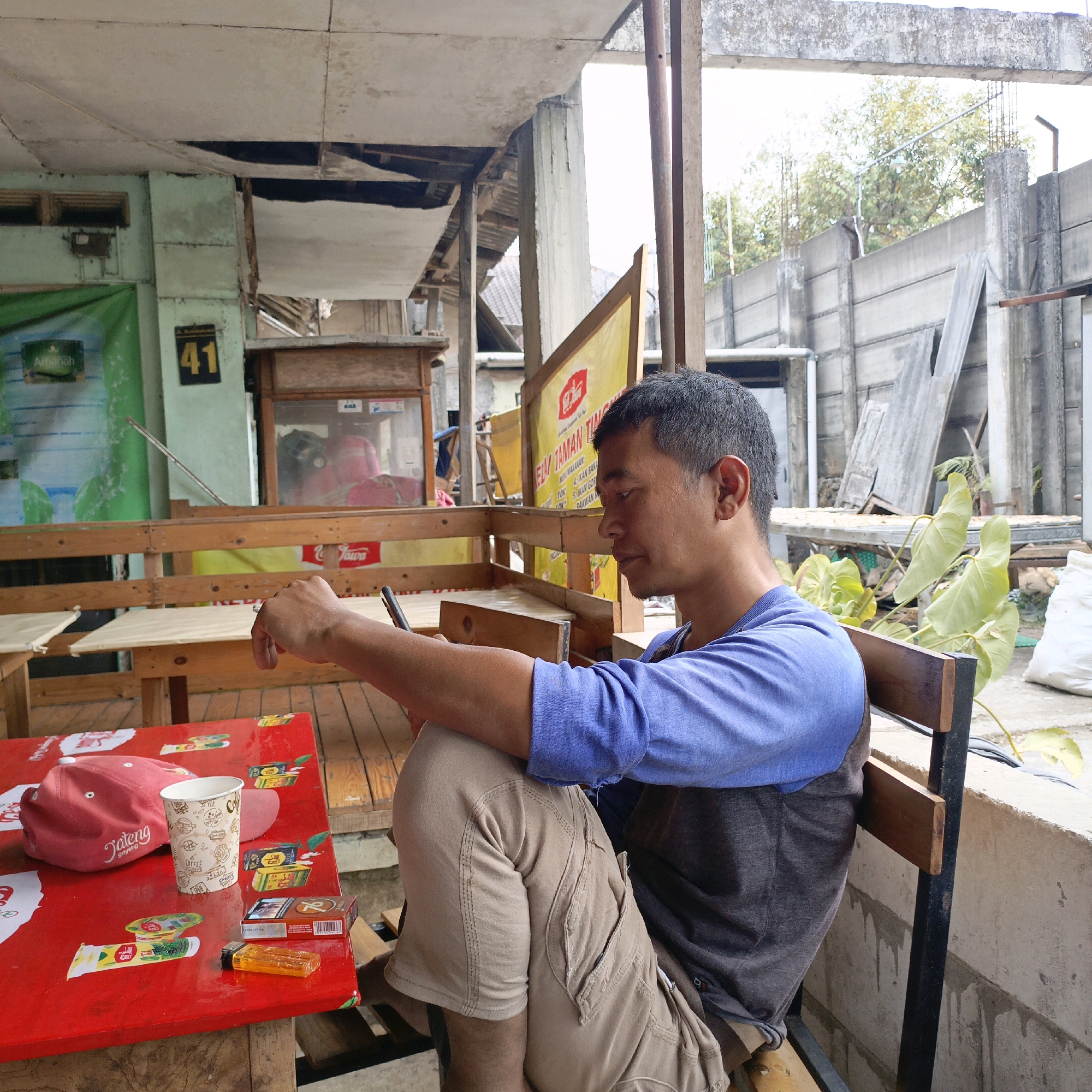 An outdoor seating area in Salatiga, Indonesia features a man sitting on a wooden bench, looking at his phone. He is wearing a blue long-sleeved shirt and tan cargo pants. On the red table in front of him are a red baseball cap, a paper cup, a cigarette pack, a lighter, and other small items. The background shows an aging building with visible structural damage, a yellow banner with text, and a concrete wall. Trees and vegetation are also visible in the background. The overall impression is that of a somewhat neglected, informal public space.