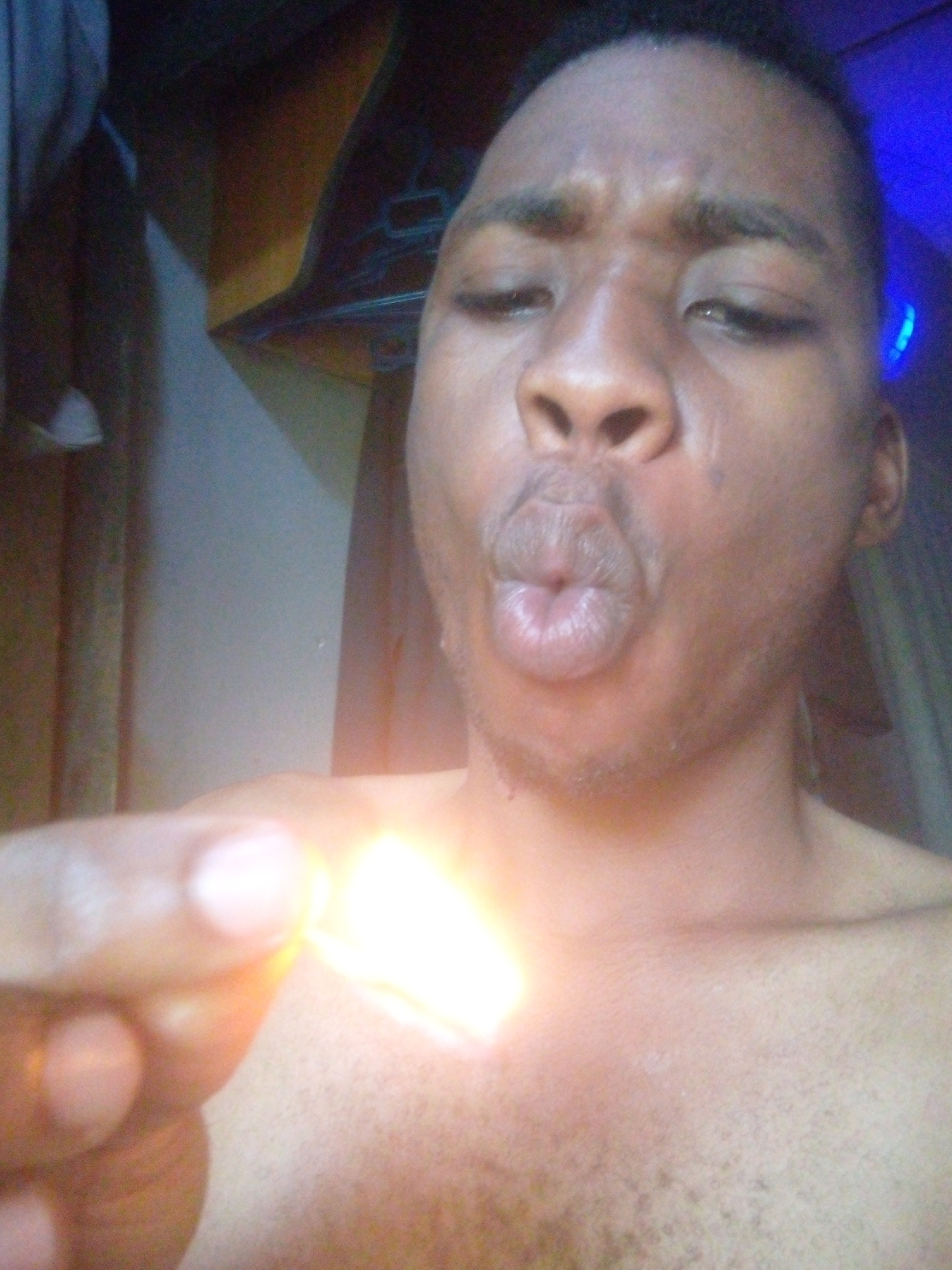 A shirtless man with dark skin, short dark hair, and facial hair is holding an object that is emitting a bright light. The man's lips are pursed into a kissing expression. The background is blurred, but includes a wall, curtain, and an overhead shelf with objects on it. The location is unspecified and unknown but the prompt states that the location is in the city of Ikorodu, Nigeria.