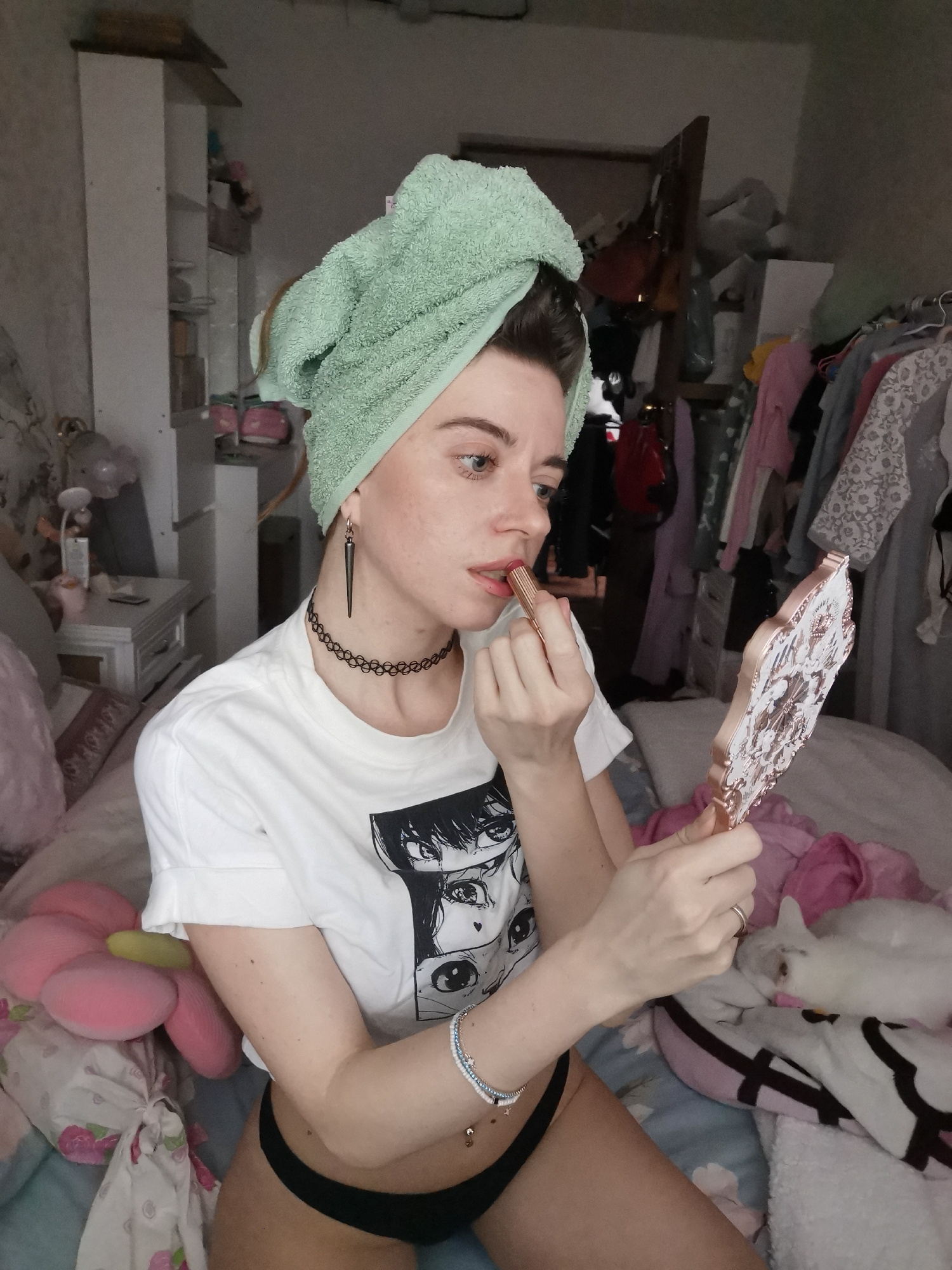 A person is captured in a medium shot indoors, likely in a bedroom, engaged in applying lipstick. They are positioned on a bed, with a light green towel wrapped around their head. The person is wearing a white t-shirt featuring a black and white graphic print depicting stylized anime or manga eyes, and black briefs.
Their left hand holds a gold-colored lipstick tube to their mouth, while their right hand holds an ornate, rose gold-framed handheld mirror, reflecting an unseen part of the room. Notable accessories include a black choker necklace, a long, dark, pointed earring on the left ear, a bracelet on the right wrist, a ring on the left ring finger, and a belly button piercing. The person's expression is focused on their reflection as they apply makeup.
The background reveals a cluttered personal space. To the left, a bedside table with various items and a floral-patterned pillow are visible. To the right and behind the person, shelves, clothes hanging on racks, and bags contribute to the messy appearance of the room. A white cat is seen resting on the bed or a blanket in the lower right corner of the frame. The bedding features floral patterns. The lighting suggests daytime indoors. No discernible text is visible other than the graphic on the t-shirt.