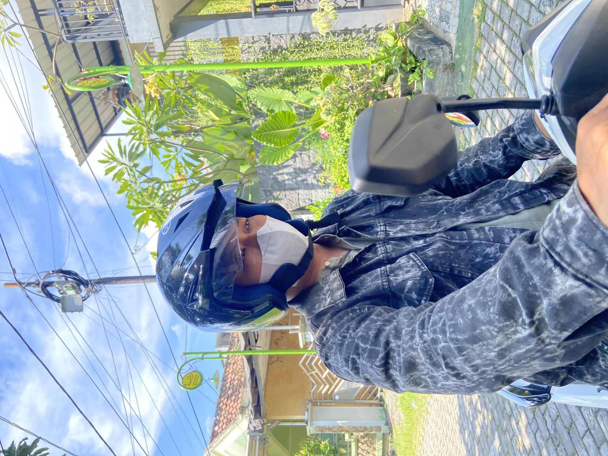 A person wearing a black motorcycle helmet and a white face mask is sitting on a parked motorcycle in Gelam, Indonesia. They are wearing a black and grey denim jacket. The background shows a street with houses, trees, and power lines against a blue sky with scattered clouds. A green sign with yellow writing is visible on a pole near a building. The ground is paved with grey rectangular stones.