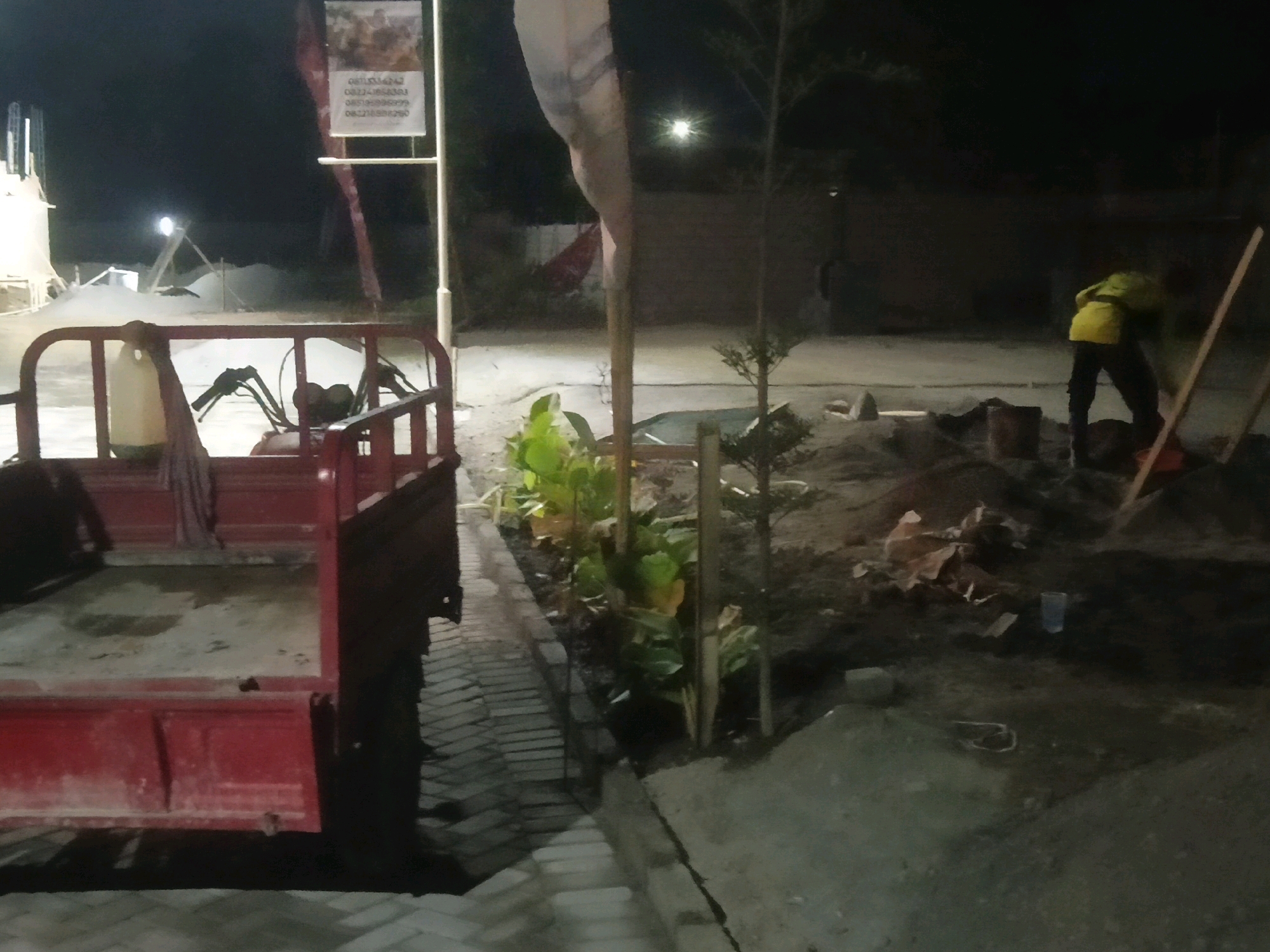 An outdoor night scene in Sendangadi, Indonesia, shows construction or landscaping work in progress. A red truck bed is prominently displayed in the foreground. In the middle ground, a person wearing a yellow jacket and dark pants is engaged in some activity involving dirt and possibly tools, near a bucket and a pile of debris. To the left of the worker, there is a small section of plants and foliage. A vertical pole with a sign at the top is visible in the background, with some text and an image on it. The ground appears to be unpaved and uneven, with piles of dirt and scattered materials. A bright light source illuminates the scene from the left, casting shadows.