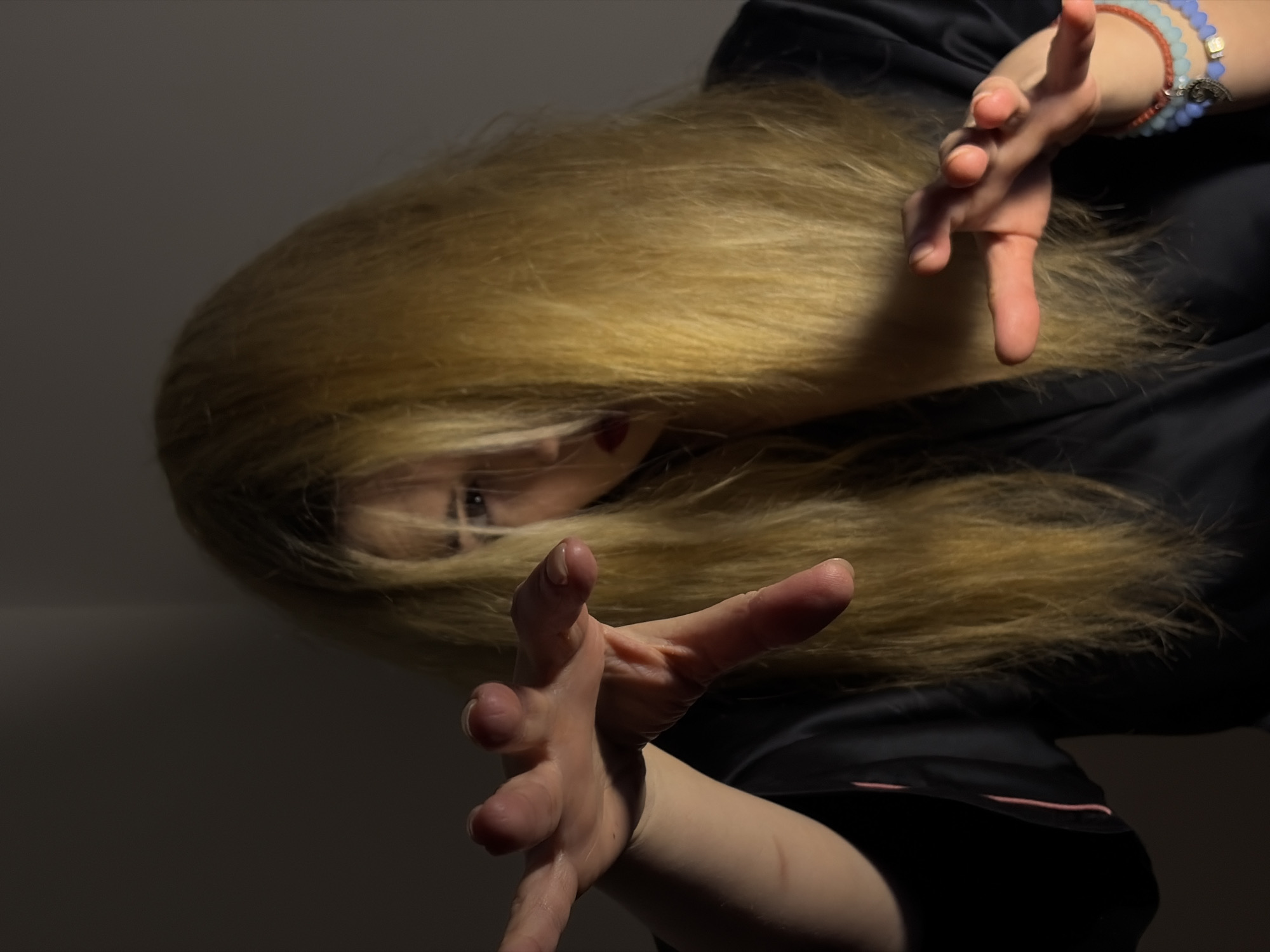A person with long, blonde hair, wearing a dark robe, is captured in a low-angle shot against a dark background. Their face is partially obscured by their hair, but one eye and red lips are visible. Both hands are raised in front of them, fingers outstretched, as if reaching towards the viewer. A multi-colored beaded bracelet is visible on the right wrist. The lighting is dim and dramatic, creating shadows across the face and hands. The city of Budva, Montenegro, is not discernible from the provided image and therefore does not factor into the description.