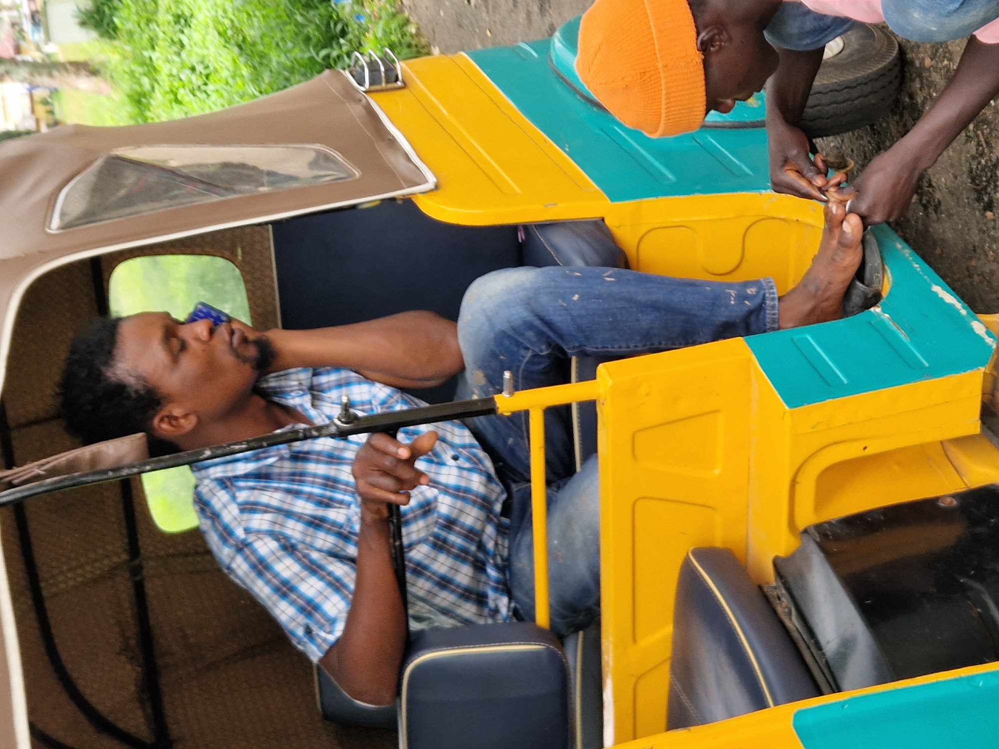 A man wearing a blue and white checkered shirt and blue jeans is sitting in the driver's seat of a yellow and teal auto-rickshaw. He has his right hand on his chin and is looking down. Another person, wearing an orange knitted cap and a pink shirt, is kneeling beside the auto-rickshaw and appears to be cutting the toenails of the man in the driver's seat. The auto-rickshaw has a brown fabric top and a dark blue upholstered seat. The background shows lush green vegetation, suggesting an outdoor setting. The image is a close-up shot of the two individuals and the auto-rickshaw. The scene suggests a moment of personal grooming or care taking place in an urban or semi-urban environment. Based on the provided context, the location is identified as Jos, Nigeria.