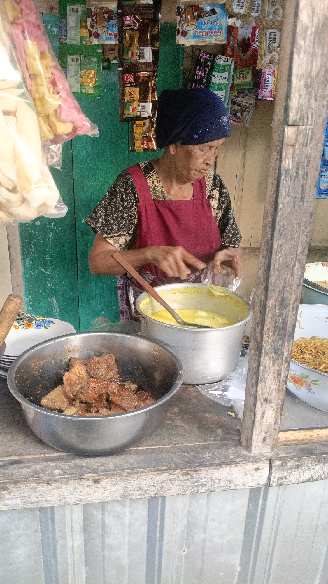 An outdoor food stall in Salatiga, Indonesia, is depicted. A woman, wearing a dark patterned shirt and a maroon apron, stands behind a wooden counter. She has a dark blue headscarf. Her hands are engaged in the action of handling a plastic bag, which she holds over a large metal pot filled with a yellow liquid. A wooden ladle rests in the pot. To the left, a silver bowl contains pieces of cooked meat. To the right, a shallow white bowl with a floral pattern holds a pile of cooked noodles. Behind the woman, shelves are stocked with various packaged goods, including snacks and beverages. The stall has a rustic wooden structure and a green wooden wall behind it. The foreground shows a corrugated metal surface. The lighting suggests daytime.