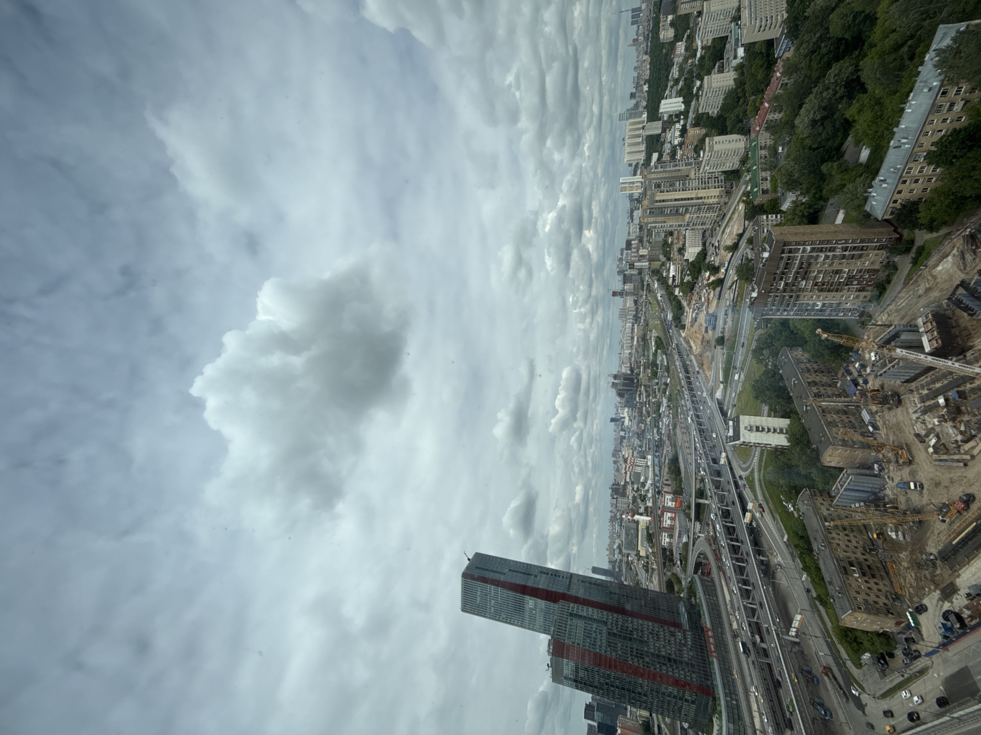 This high-angle, wide-shot photograph captures a vast urban landscape in Khimki, Russia, under an overcast sky. The image is oriented vertically but depicts a horizontal city vista.
The sky is filled with dense, light grey to white clouds, suggesting an overcast day. In the foreground, on the left, a prominent, tall, modern skyscraper, dark in color with distinctive red vertical bands, stands out. Adjacent to it, a complex network of multi-lane highways and elevated road sections stretches across the mid-ground, bustling with vehicle traffic.
To the right of the highway, the cityscape features a mix of older and newer buildings. Several multi-story residential blocks, some appearing to be Soviet-era structures, are interspersed with areas of dense green trees. Further to the right, significant construction activity is visible, characterized by multiple yellow tower cranes, excavated earth, and partially built structures. Some legible text elements include "TENK" on one crane arm, possibly "KONE" on another, and "ЭТЕНА" (ETENA) on a construction barrier.
The background reveals a sprawling city extending to the horizon, with numerous buildings and green spaces blending into the distance. The overall scene portrays a large, active metropolitan area undergoing continuous development, characterized by extensive infrastructure and diverse architectural styles under cloudy daytime conditions.