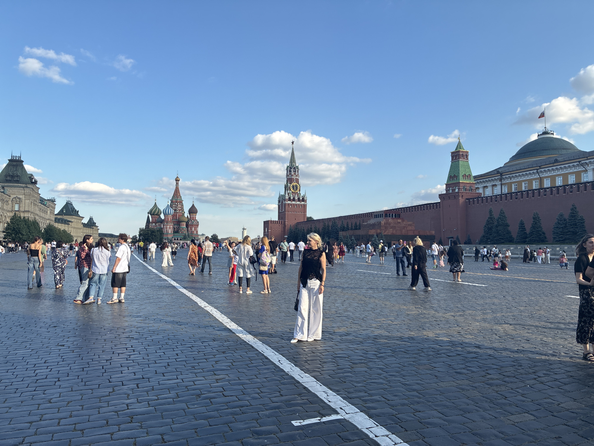 The image depicts Red Square in Moscow, Russia, on a clear, sunny day. A wide, paved area, likely the square itself, is visible in the foreground, composed of dark cobblestones. A white line is painted on the cobblestones, running from the bottom left corner towards the center of the image. Numerous people are scattered across the square, walking, standing in groups, and posing.
In the background, iconic Moscow landmarks are present. To the left, a large building with a green roof and ornate facade, possibly part of the GUM department store, is partially visible. The central focus of the background is the Spasskaya Tower, a prominent clock tower with a red brick structure and a distinctive spire topped with a red star. To the right of the Spasskaya Tower, sections of the red brick wall of the Moscow Kremlin can be seen, along with a green-roofed tower. Further to the right, a large building with a green domed roof, likely the State Historical Museum or Senate building, is visible, featuring a Russian flag flying from its apex.
The sky is a vibrant blue with scattered white clouds, indicating good weather. The lighting suggests it is daytime, with the sun casting shadows on the cobblestones and illuminating the scene. The people in the image are dressed in casual summer attire, including dresses, shirts, pants, and shorts. Some individuals are looking at their phones, while others are engaged in conversation or observing the surroundings. The overall atmosphere appears to be one of tourism and public gathering in a significant historical and cultural location.