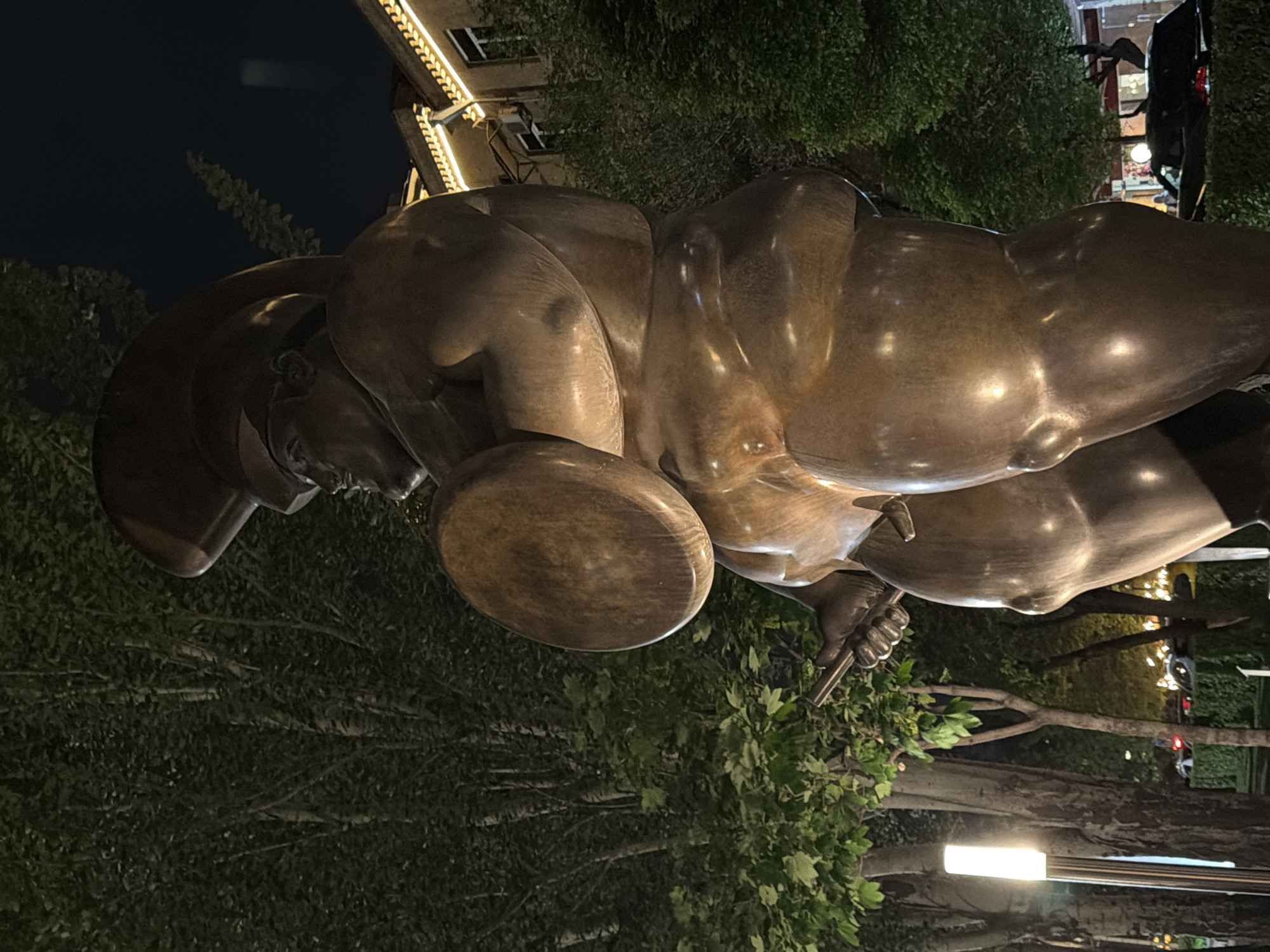 This image shows a bronze statue of a gladiator, likely a historical or mythological figure, with a helmet and shield, placed outdoors in Yerevan, Armenia. The statue is positioned against a backdrop of trees and possibly other greenery. In the background, there are buildings, streetlights, and some evidence of urban activity, possibly a passing car. The lighting is a combination of ambient and artificial, indicating it might be nighttime or dusk. The statue itself appears to be the primary subject, with surrounding elements acting as contextual details.