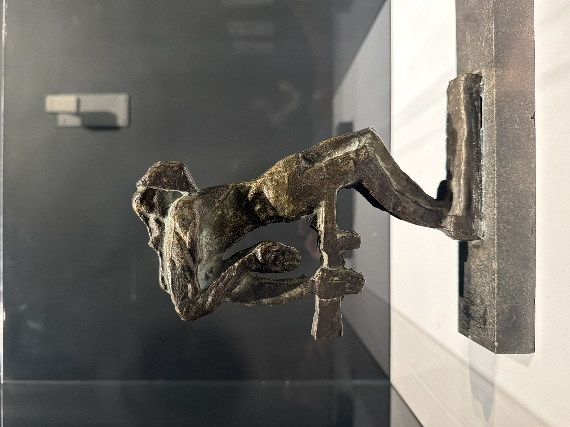 A bronze sculpture of a man on a cross is on display in the Kolodzei Art Foundation in Moscow, Russia. The sculpture is attached to a rectangular mounting bracket on a white wall in front of a glossy black wall that reflects the sculpture.
