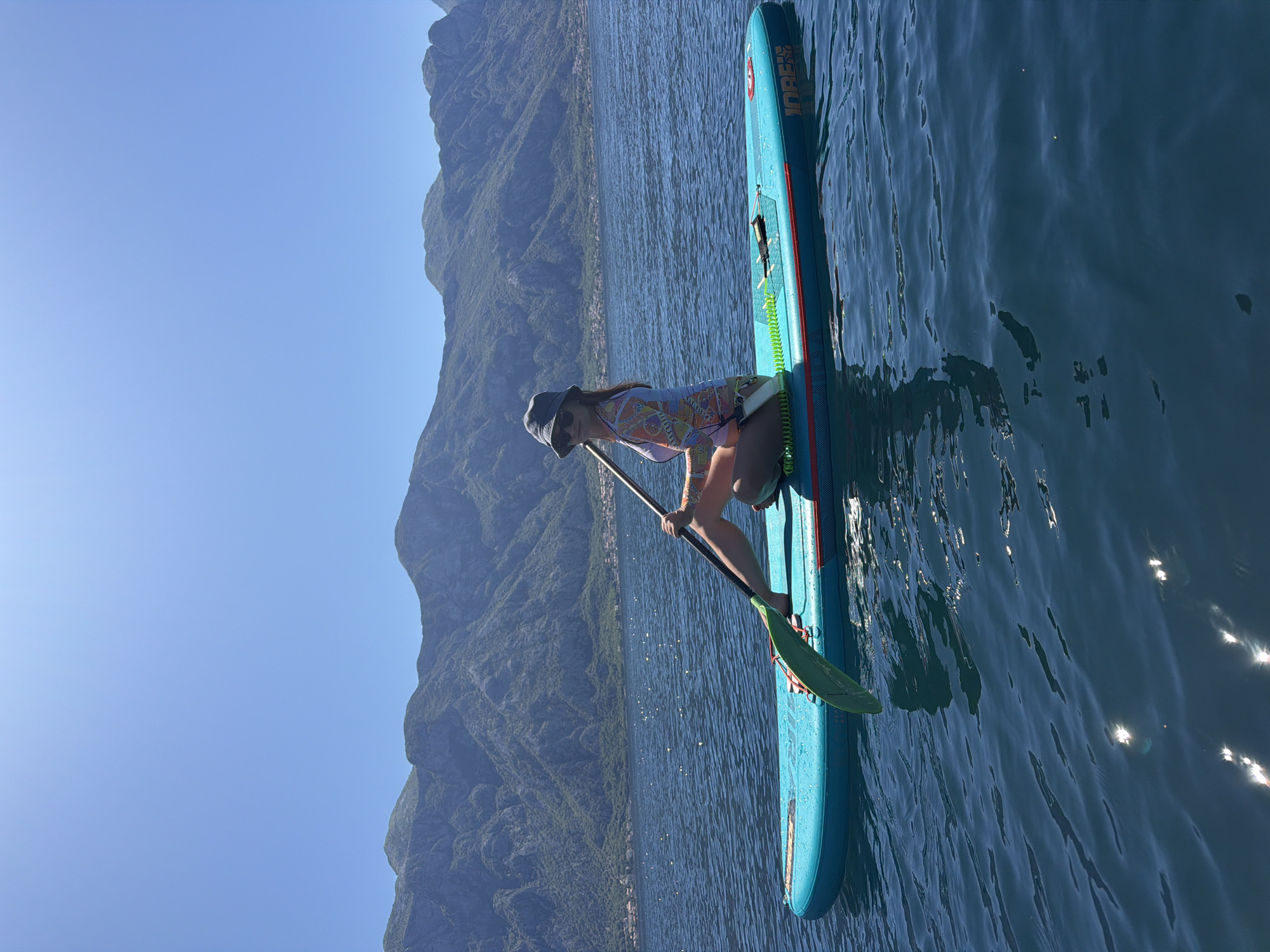 A woman is stand-up paddleboarding on the Bay of Kotor, in the vicinity of Stoliv, Montenegro. She is wearing a long-sleeved rash guard, a hat, and sunglasses. She is holding a paddle in the water. The paddleboard is blue. In the background is a mountain range and a clear blue sky. The water is a deep blue color with sunlight reflecting off the surface.